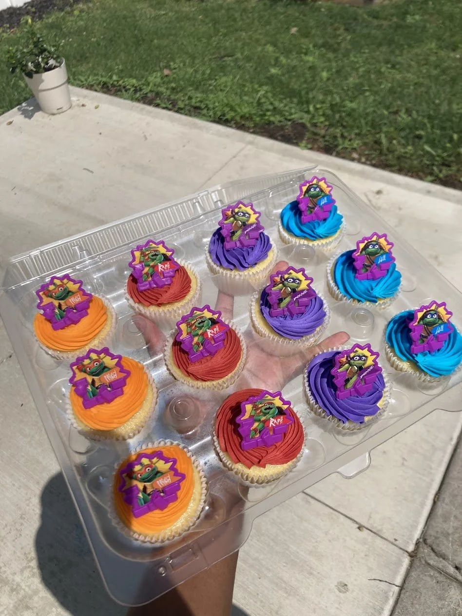 A hand holding a clear plastic container with cupcakes decorated with characters from the Teenage Mutant Ninja Turtles in orange, red, purple, and blue frosting.