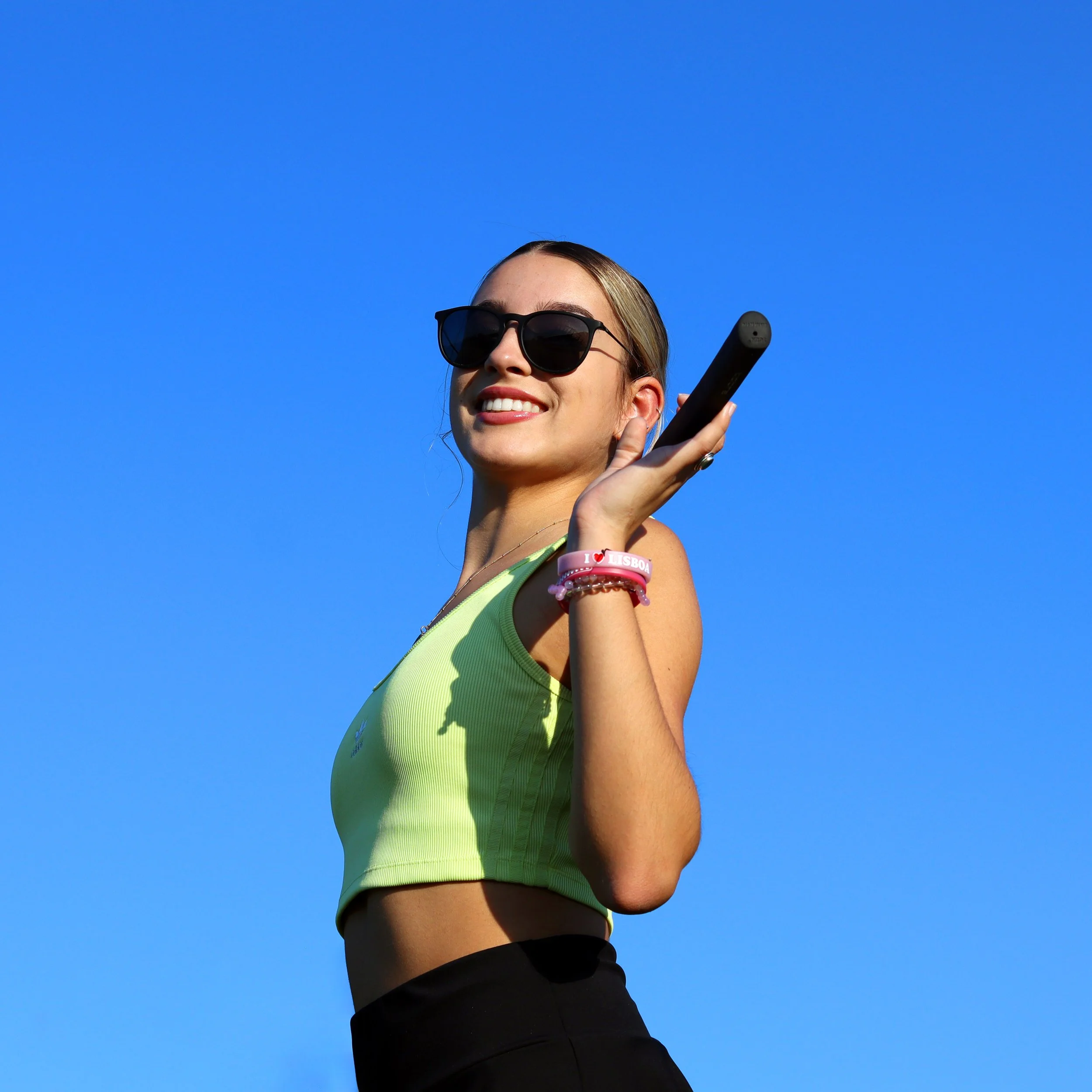 Young woman wearing sunglasses, smiling, holding a tennis racket over her shoulder against a clear blue sky.