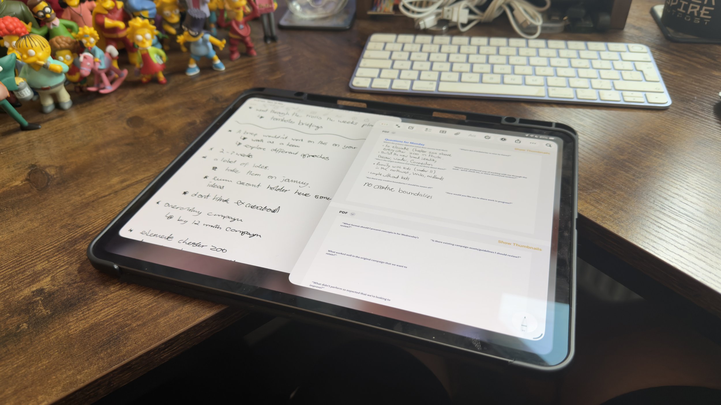 A tablet on a wooden desk displaying handwritten notes and a PDF document, with a white wireless keyboard and a collection of small figurines of cartoon characters nearby.