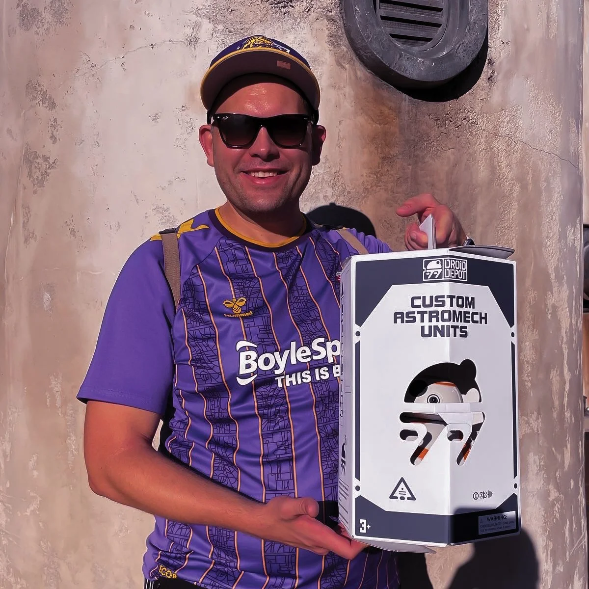 Matt Reaney wearing a purple and yellow sports jersey, sunglasses, and a cap, holding a white box labeled 'Custom Astromech Units' with a small dome-shaped robot inside visible through a cutout.