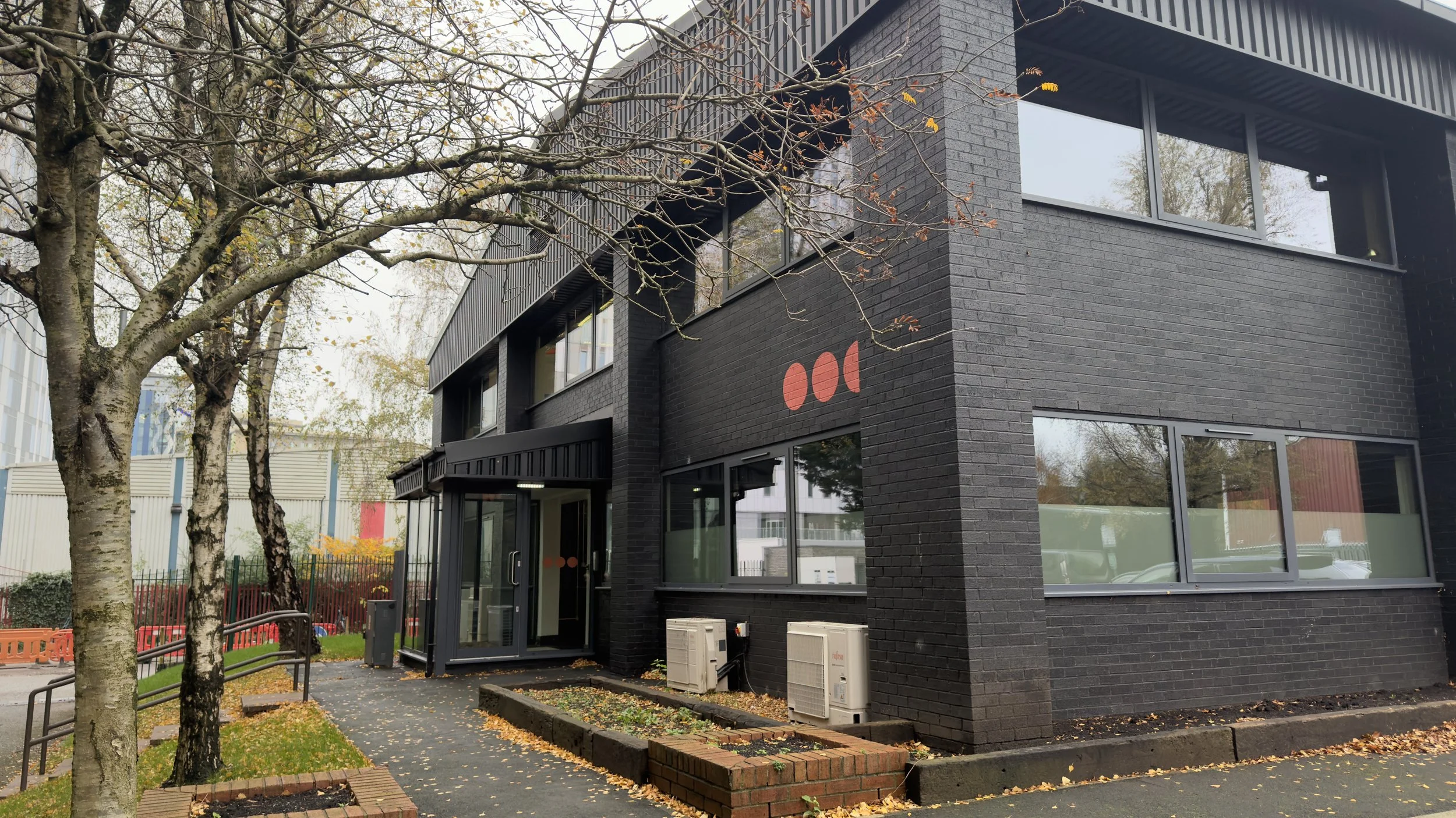 Modern black brick building with large windows, surrounded by trees with fallen autumn leaves on the ground.