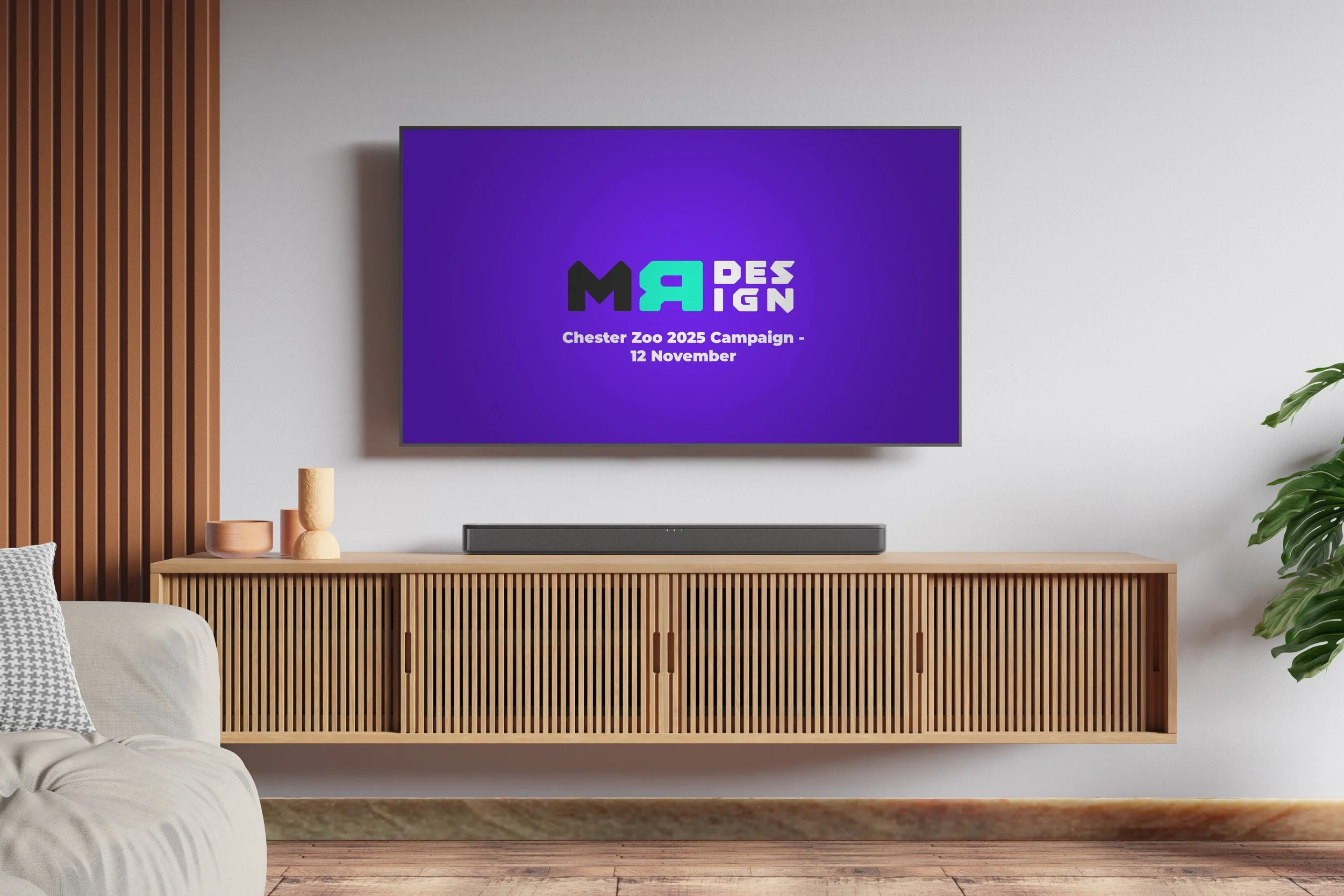 Modern living room with a mounted TV displaying a purple screen with colorful text, a wooden media console, a soundbar, decorative vases, a corner of a sofa with a patterned pillow, and a large green plant, with a wood-paneled wall on the left.