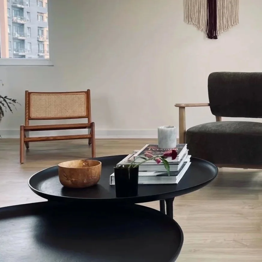 A modern living room with a black coffee table holding a wooden bowl, a black vase with pink flowers, a stack of magazines, and a white candle. There is a wooden chair with a woven back, a dark upholstered sofa, and a wall hanging with fringes, with