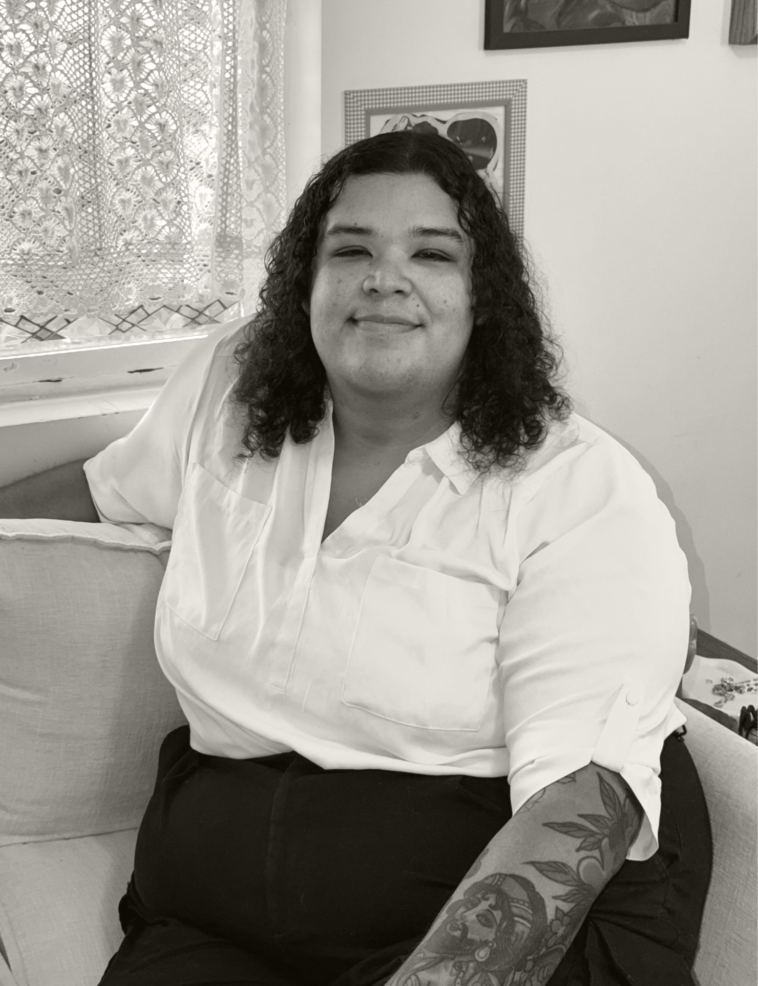 Aaron Roladan sitting on a couch, smiling, with curly hair and wearing a white shirt and dark pants, with a tattooed arm resting on her lap.