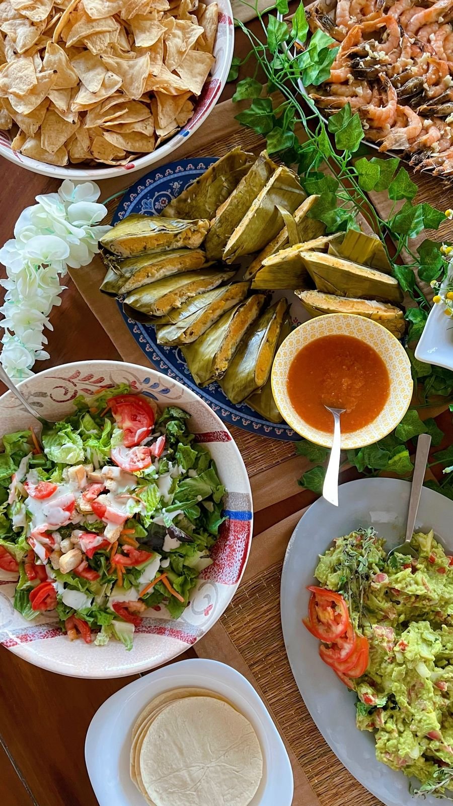 A table with assorted dishes including tortilla chips, tamales, shrimp, salad with dressing, guacamole with tomatoes, tortillas, and a bowl of salsa.