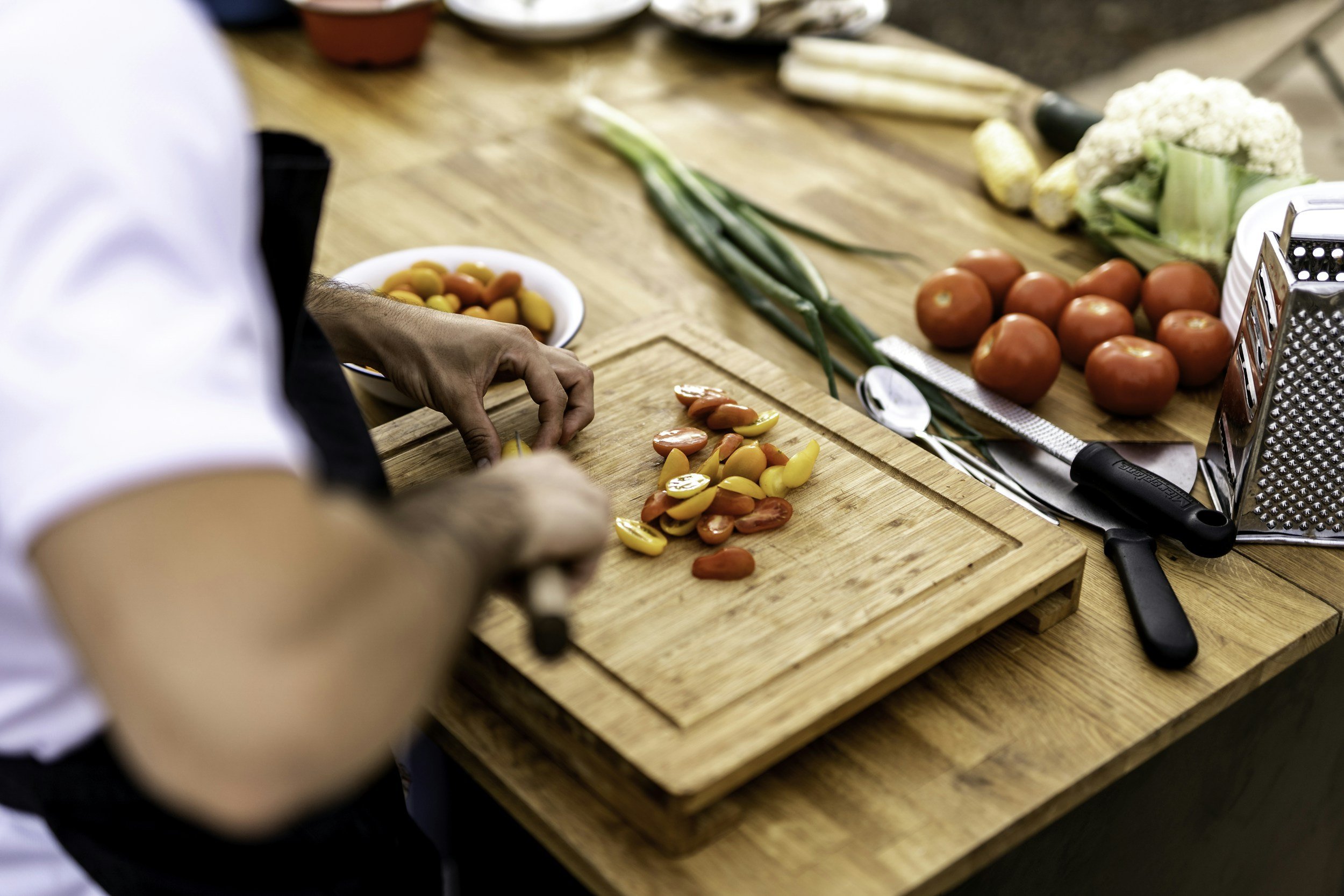 Person slicing cherry tomatoes on a wooden cutting board, surrounded by kitchen utensils, onions, and whole tomatoes.