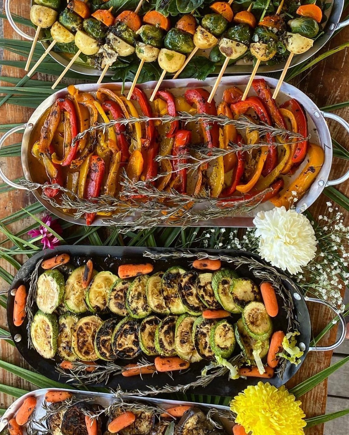 An array of grilled vegetables presented on trays. The top tray features skewered vegetables including potatoes, sweet potatoes, and Brussels sprouts on a bed of leaves. The middle tray showcases grilled red and yellow bell peppers garnished with spr