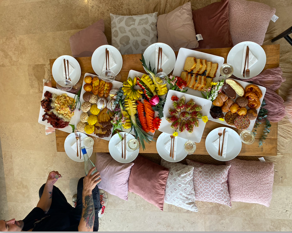 Table with assorted breakfast foods, fruits, pastries, bread, and plates, surrounded by cushions in Tulum.
