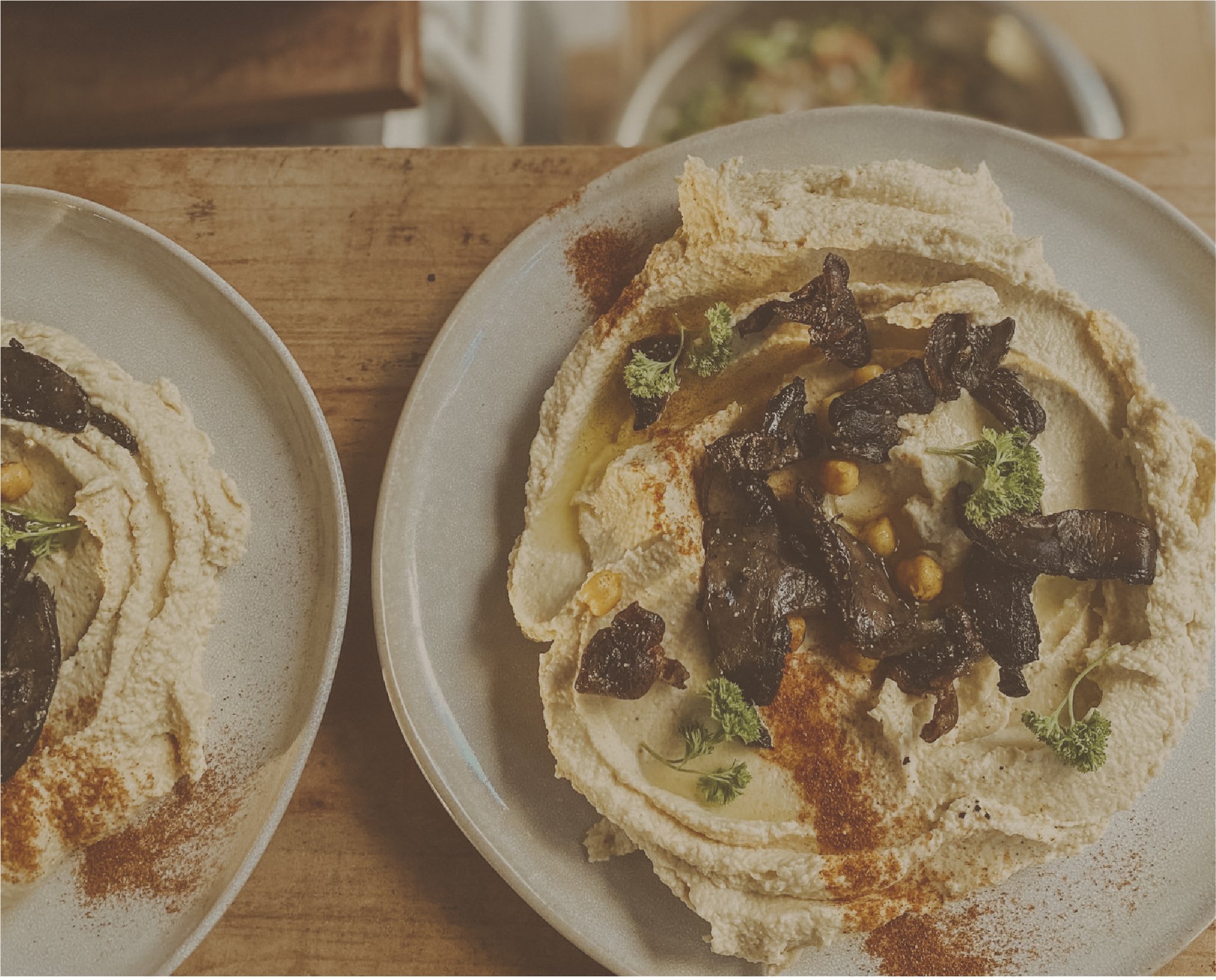 Plates of hummus topped with roasted mushrooms, chickpeas, parsley, and paprika on a wooden table.