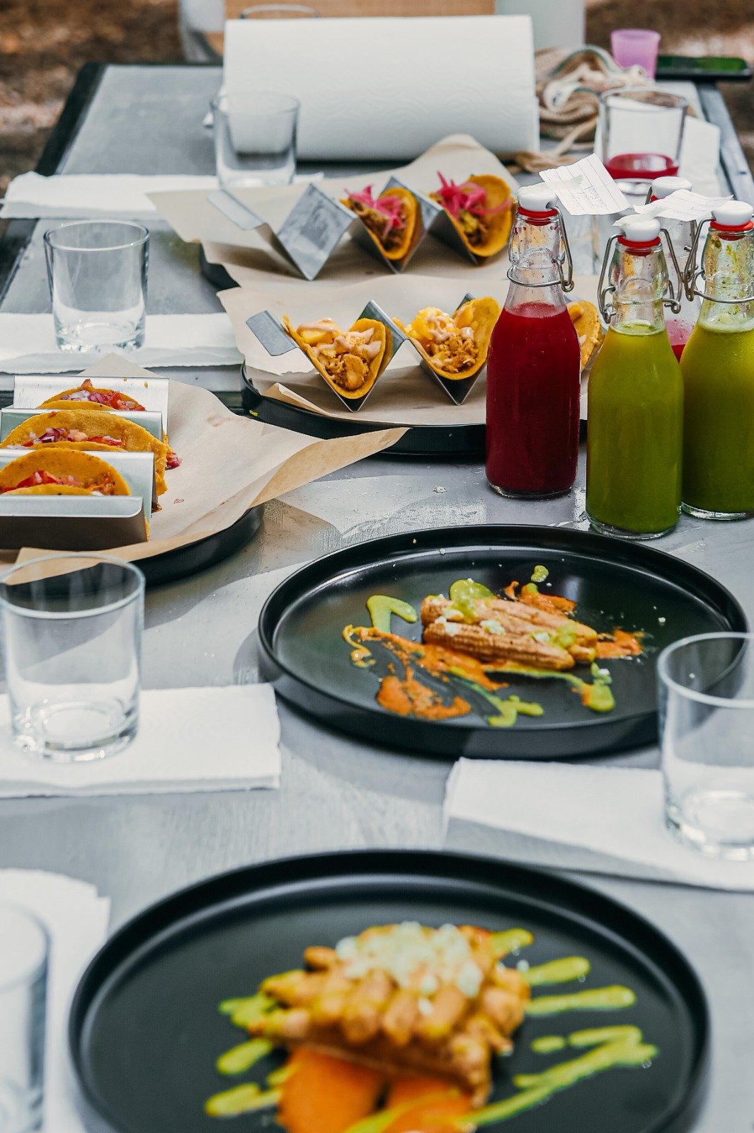 Assorted tacos and grilled dishes on a table with glass bottles of colorful beverages and empty glasses.