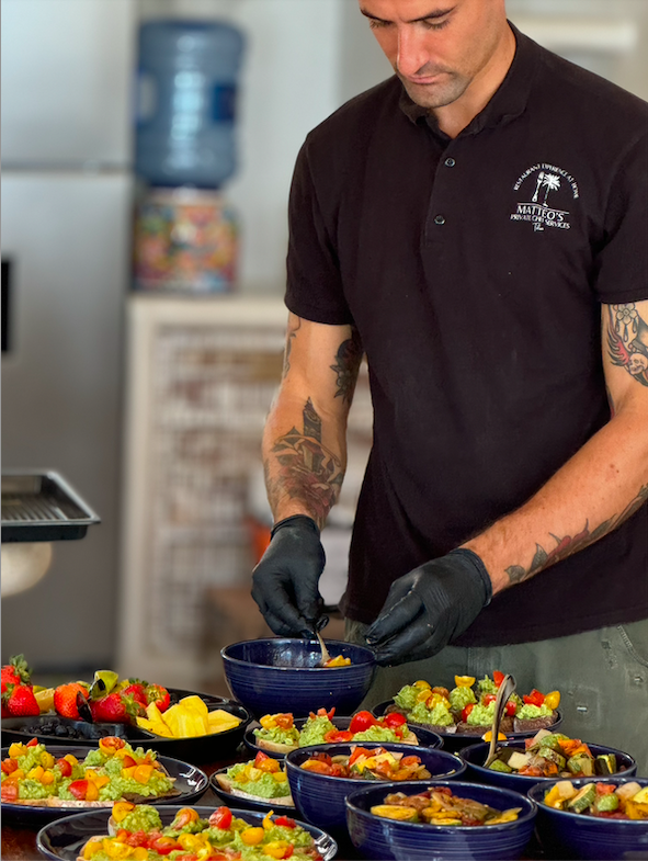 A person preparing colorful salad dishes on a table, wearing black gloves and a dark polo shirt. The salads contain a variety of fruits and vegetables.