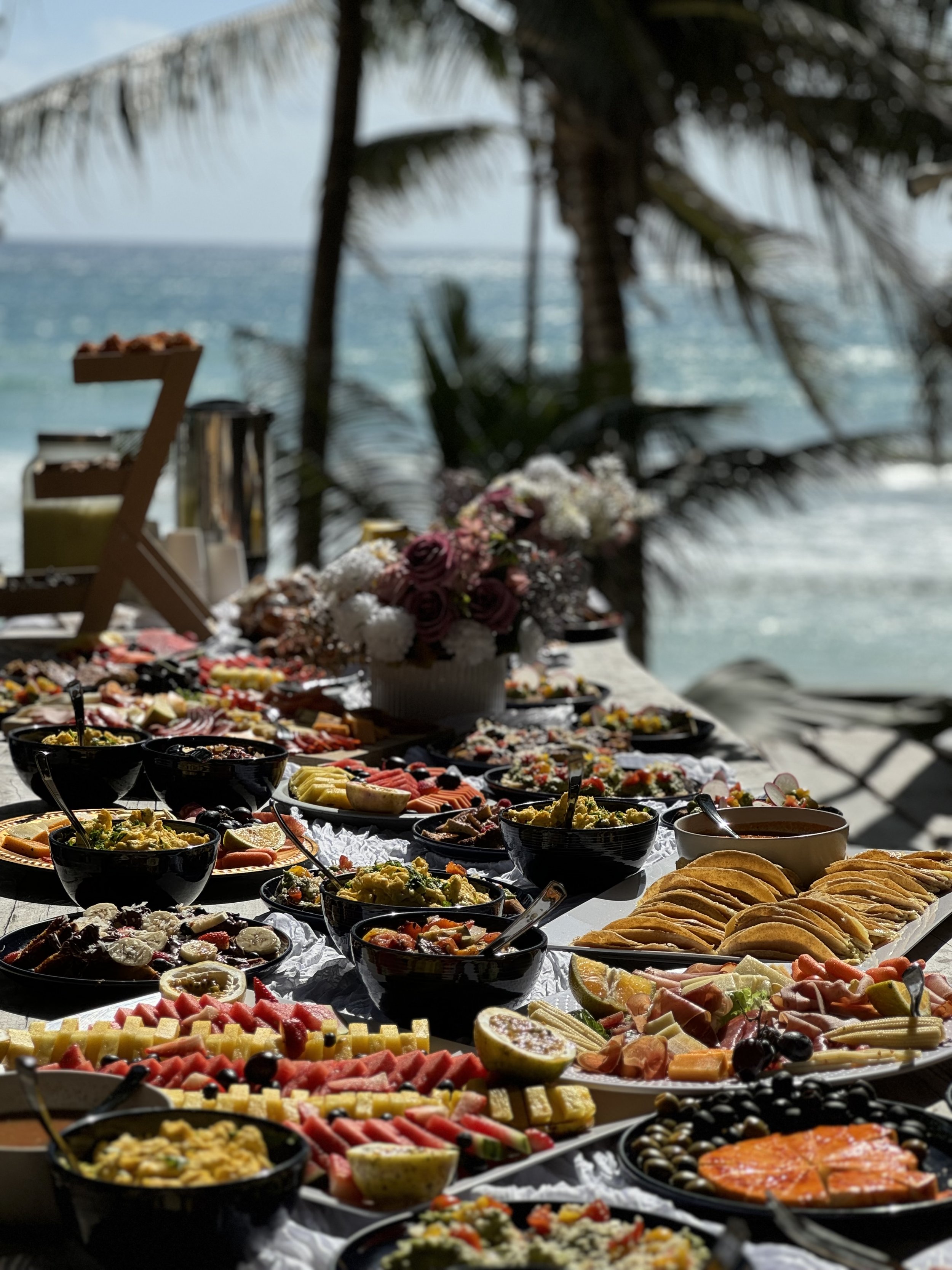 A tropical buffet spread with various dishes, fruits, and desserts set up outdoors near the ocean, surrounded by palm trees.