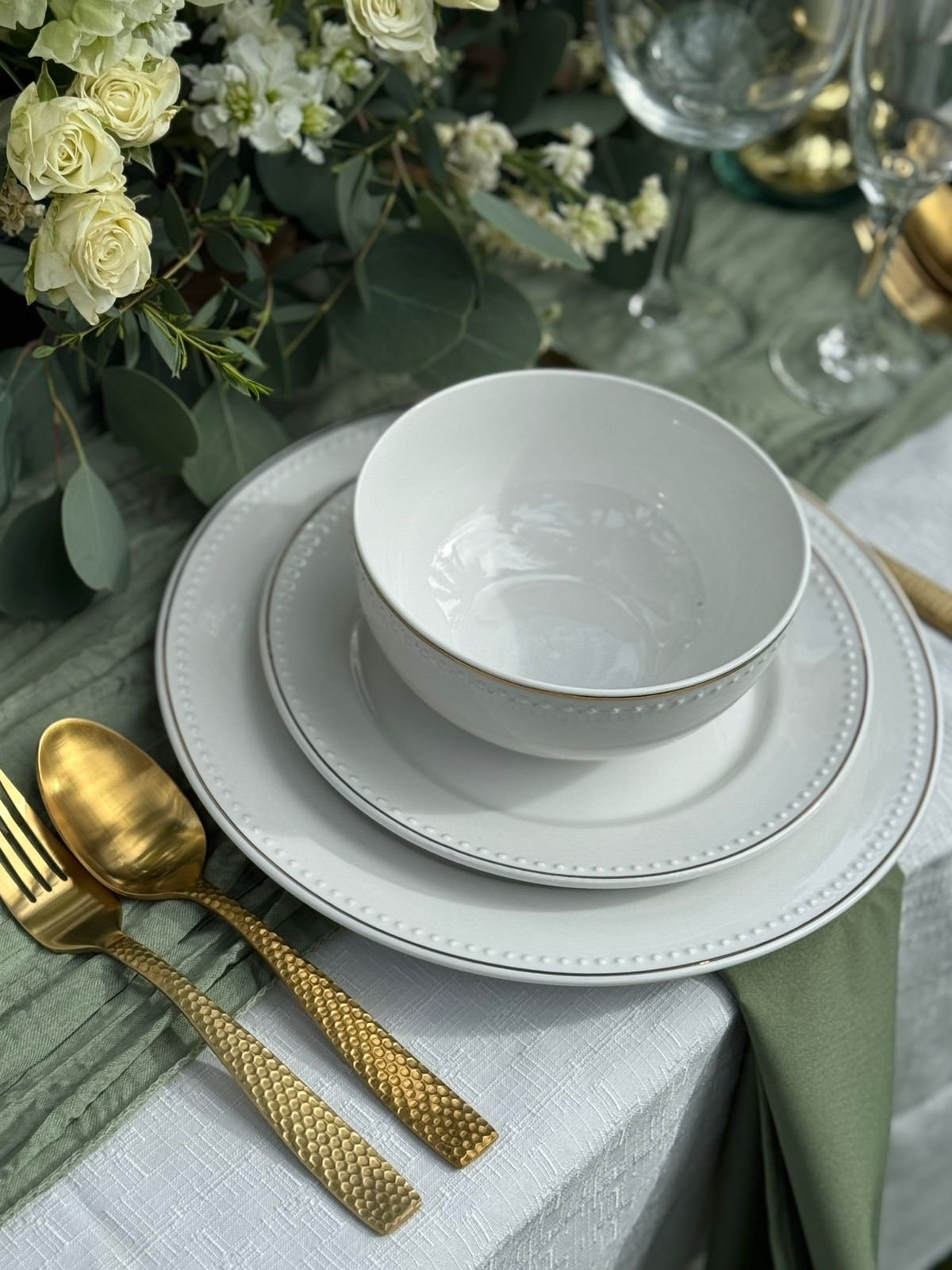 Elegant table setting with white plates, a bowl, gold cutlery, and a green napkin. Floral arrangement with white roses and greenery. White textured tablecloth and green runner.