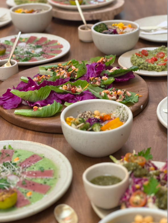 A variety of colorful dishes on a wooden table, including leaf-wrapped appetizers on a serving board, green salad with dressing in bowls, and a plate with sliced meat drizzled with a green sauce.