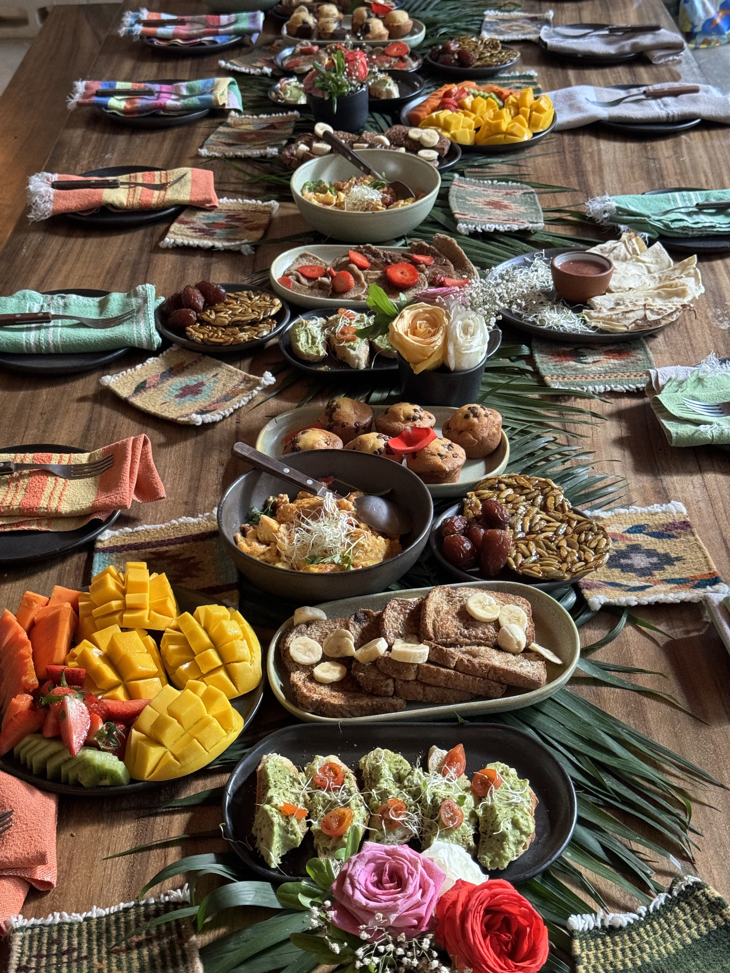 A rustic table setting with a variety of breakfast foods and colorful cloth napkins. Dishes include sliced mangoes, avocados, berries, sliced toast with banana, assorted muffins, nuts, sliced vegetables, and small bowls of spreads. Decorative element