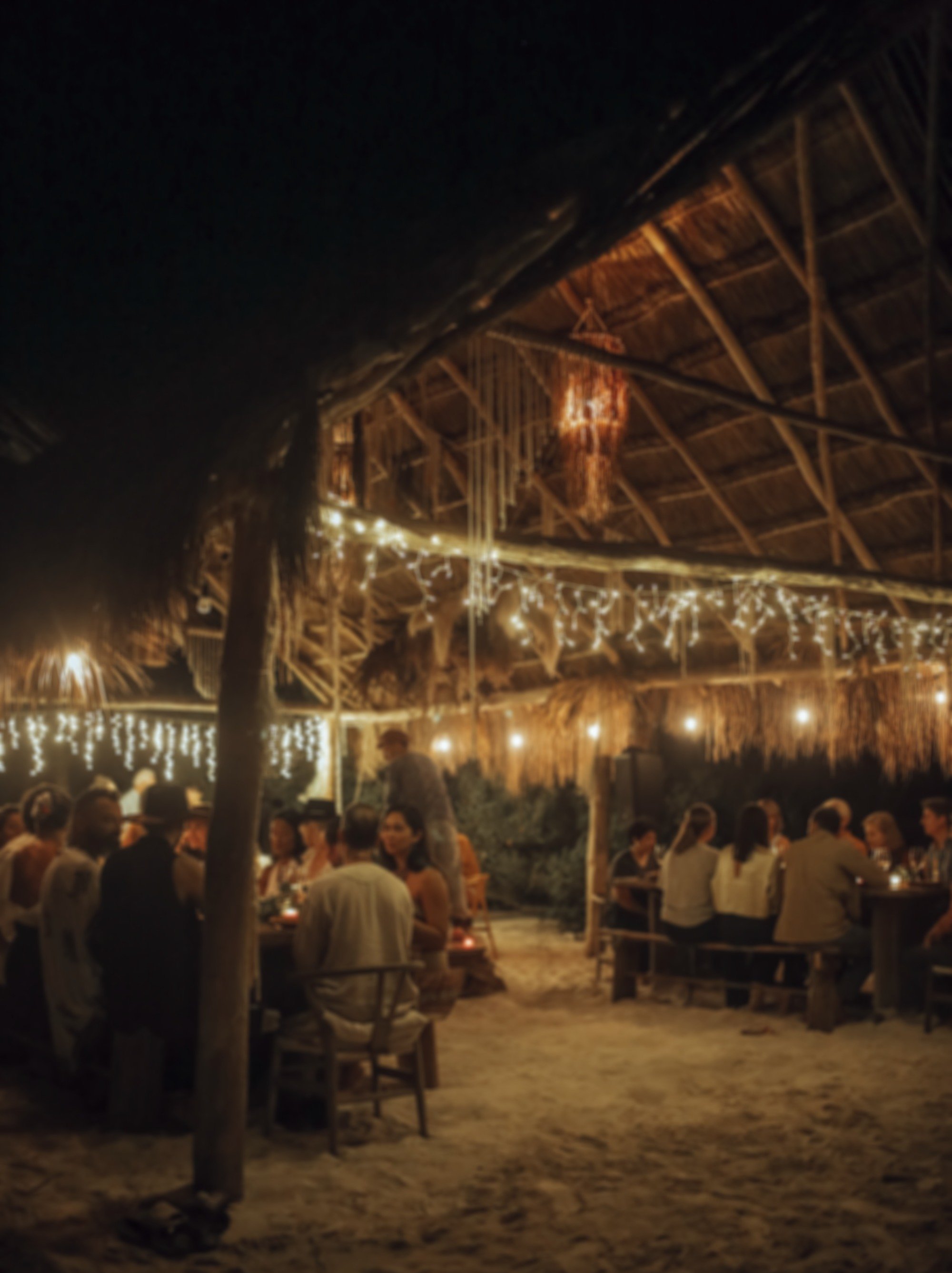 Beachside private dining experience at night under a thatched roof in Tulum with string lights, people sitting around tables, sand floor.