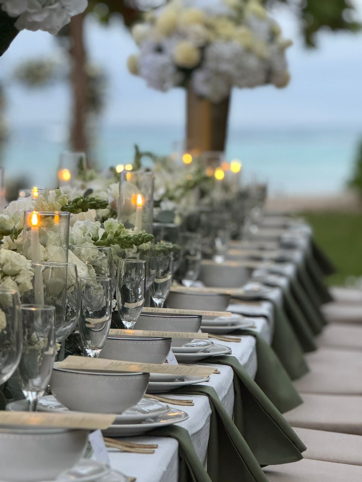Elegant outdoor dining table setting with white tablecloths, green napkins, glassware, candles, and white floral arrangements in a beachfront location.