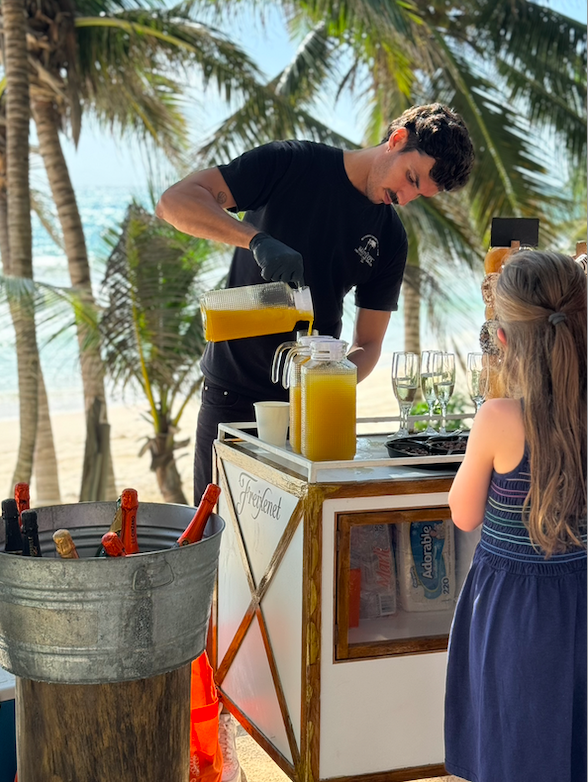 A man pouring orange juice into a pitcher at a beachside stand with palm trees in the background, a cooler with beverages, and a girl watching.