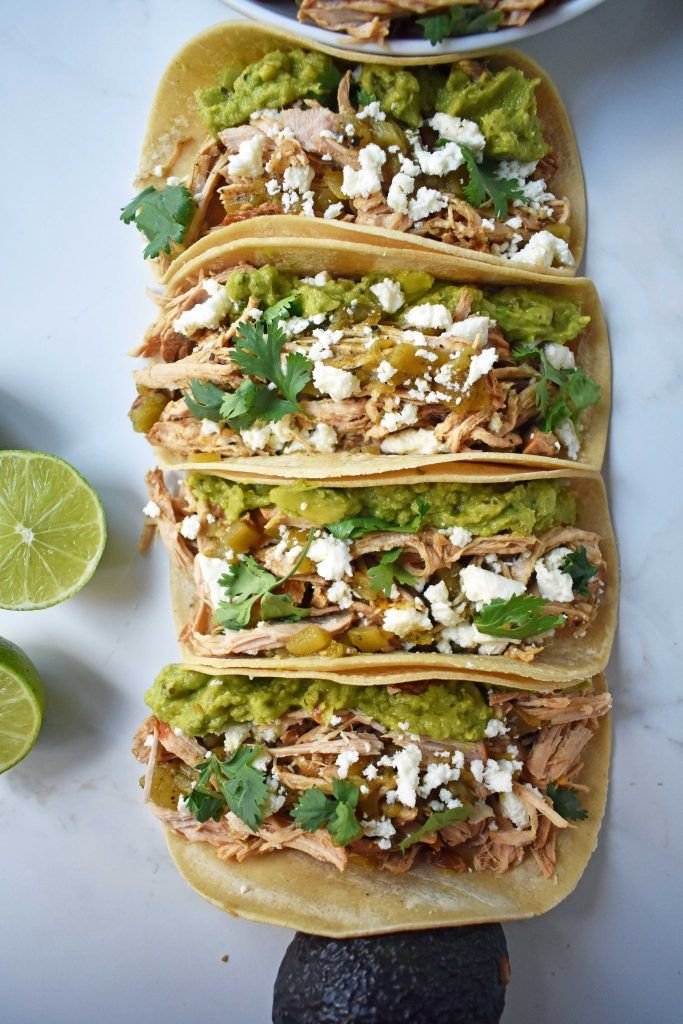 Soft tortilla tacos filled with shredded chicken, guacamole, crumbled cheese, corn, and cilantro, with lime halves and an avocado beside them on a light surface.