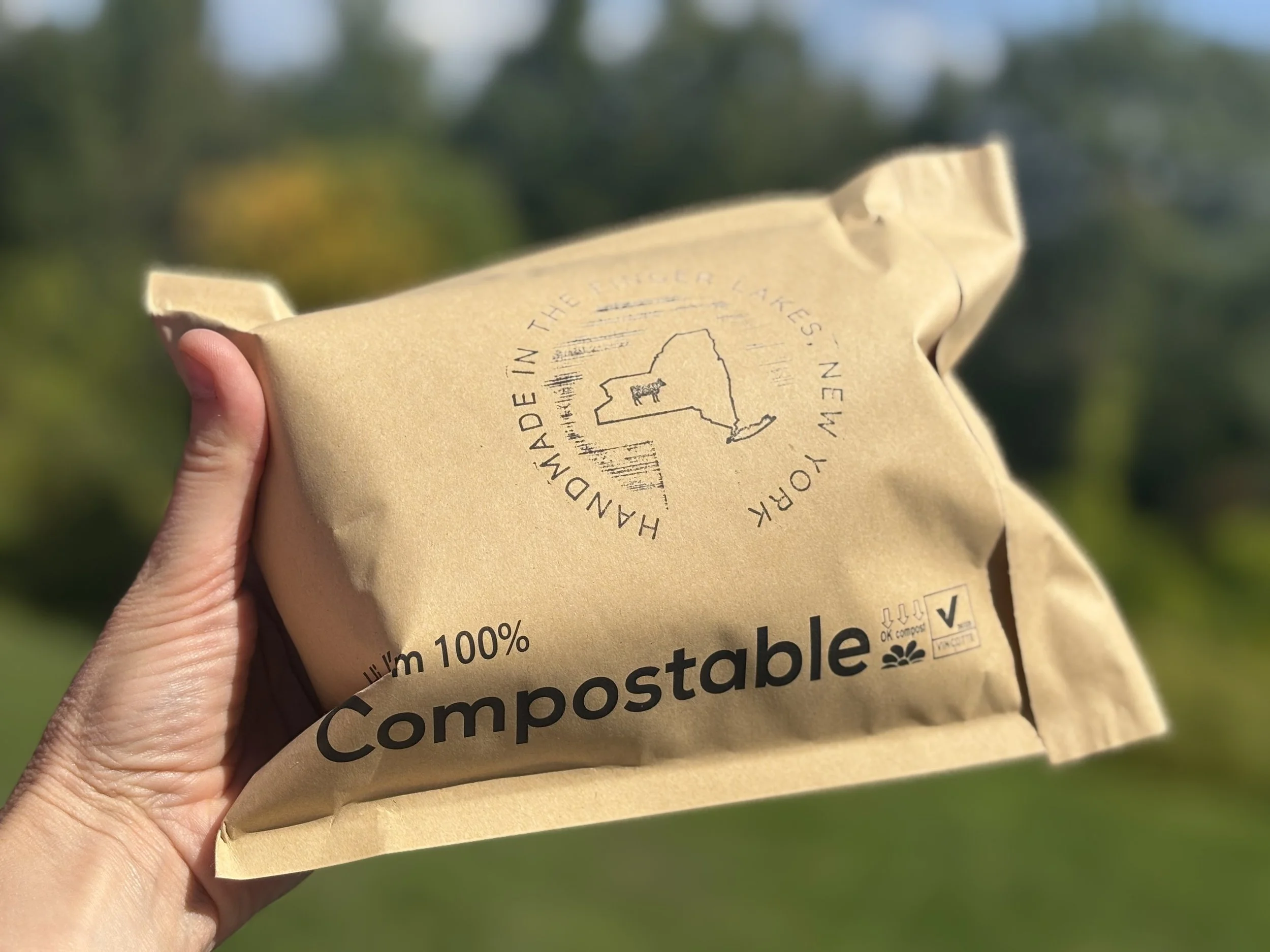 A hand holding a brown paper bag labeled '100% Compostable' with a logo of New York and a circular text stating 'Handmade in the Finger Lakes, New York, USA.' The bag is outdoors with a blurred green and blue background.