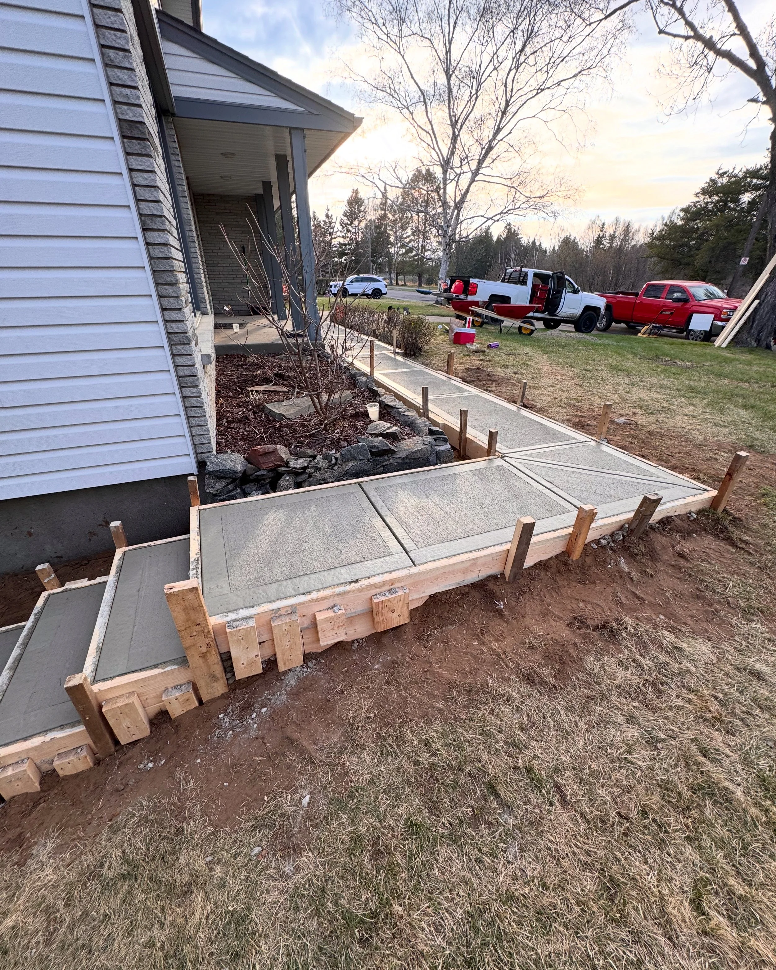 Front porch with new concrete sidewalk being installed, wooden forms and stakes in place, residential neighborhood with parked trucks and trees in the background.