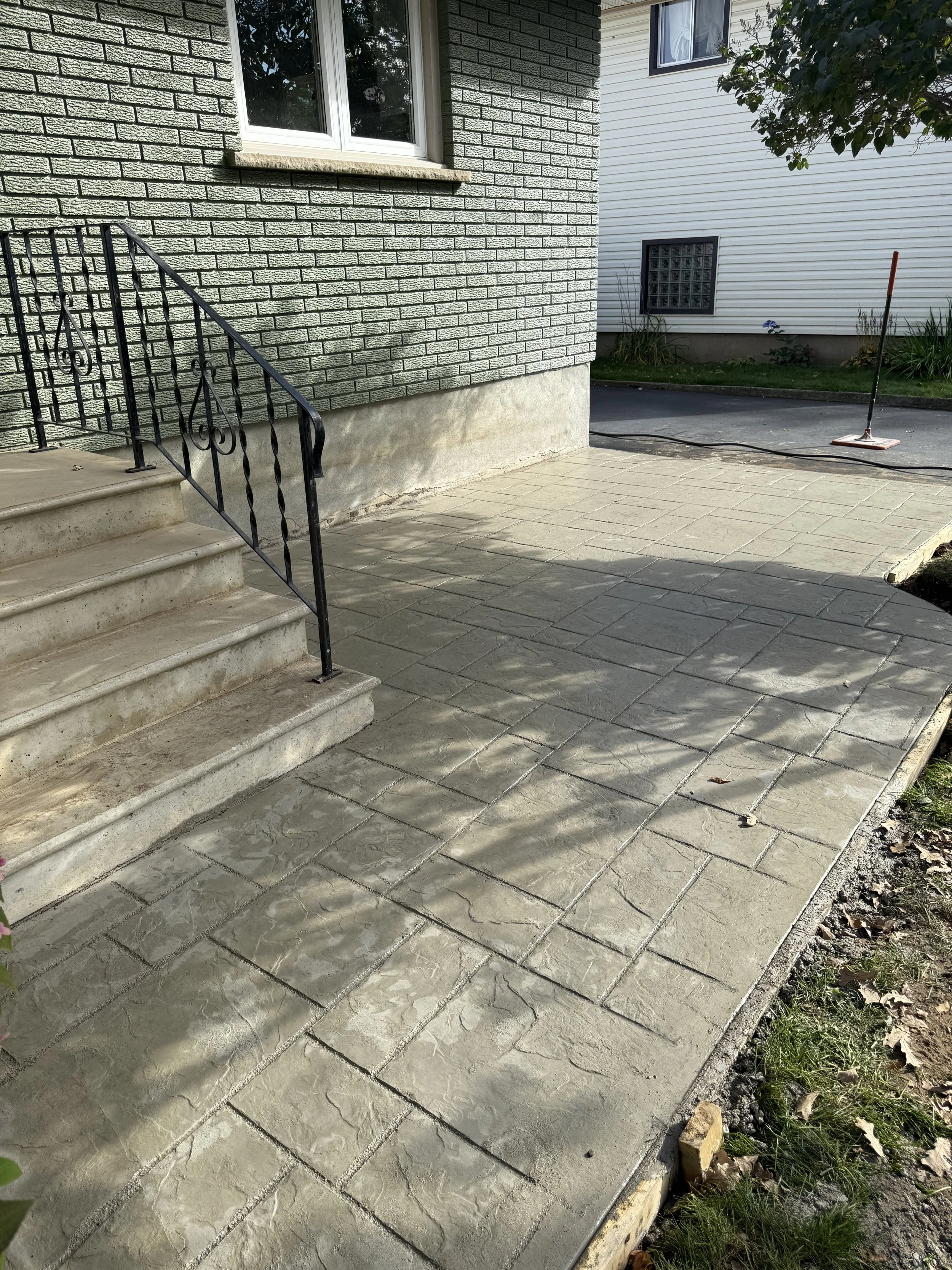 Newly poured concrete sidewalk with stamped interlock stone pattern, adjacent to a house with brick siding, black railing, and stairs. in Thunder Bay, Ontario