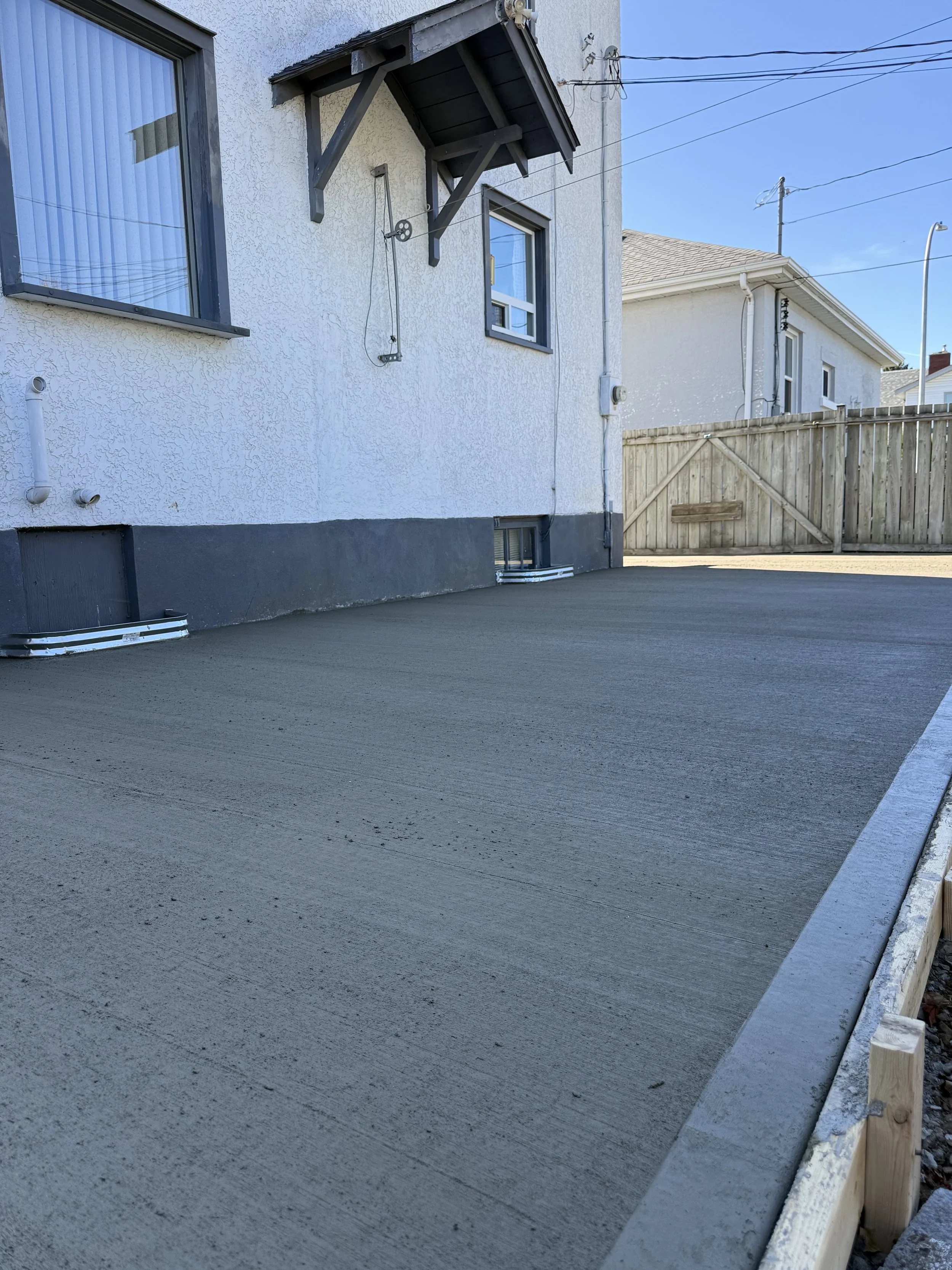 Concrete patio being freshly poured outside a house with white exterior walls, two windows, and a wooden fence in the background. Thunder Bay, Ontario