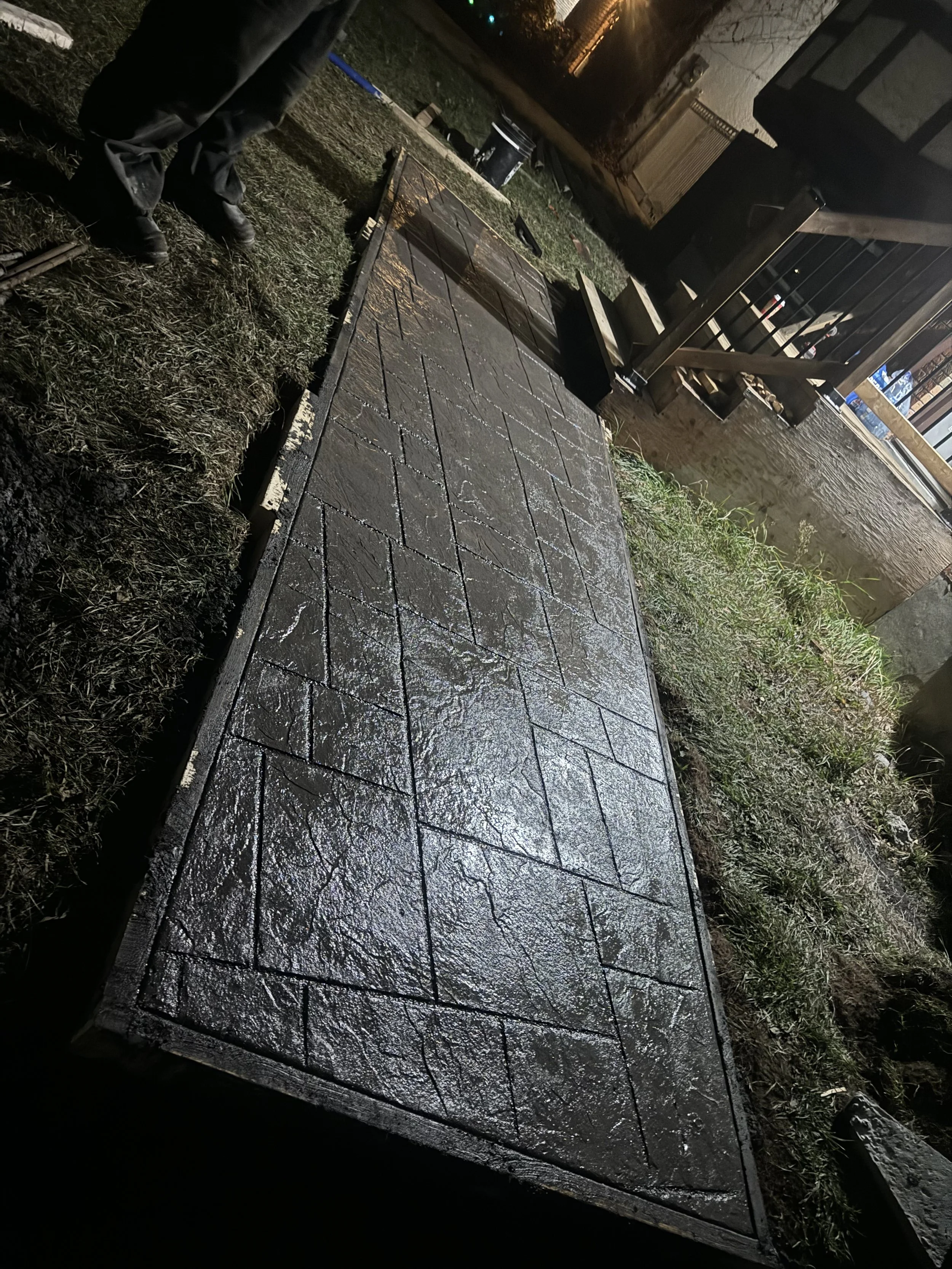 A person laying wet concrete paving stones on a sidewalk at night, with visible tools and construction materials nearby.