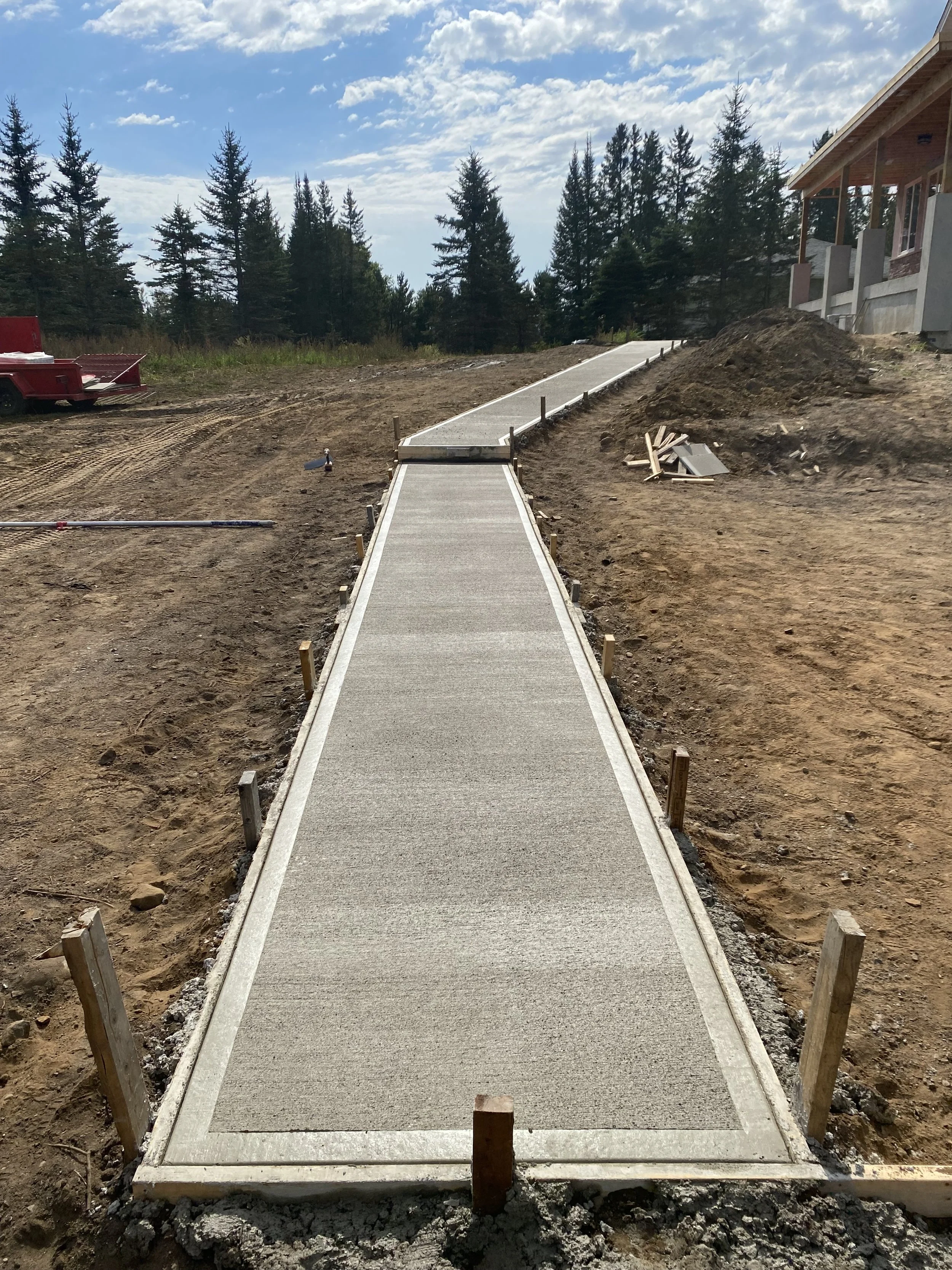 A newly constructed concrete sidewalk with wooden stakes along its edges, leading up a slight hill in an outdoor construction site surrounded by dirt, trees, and a partially built house.