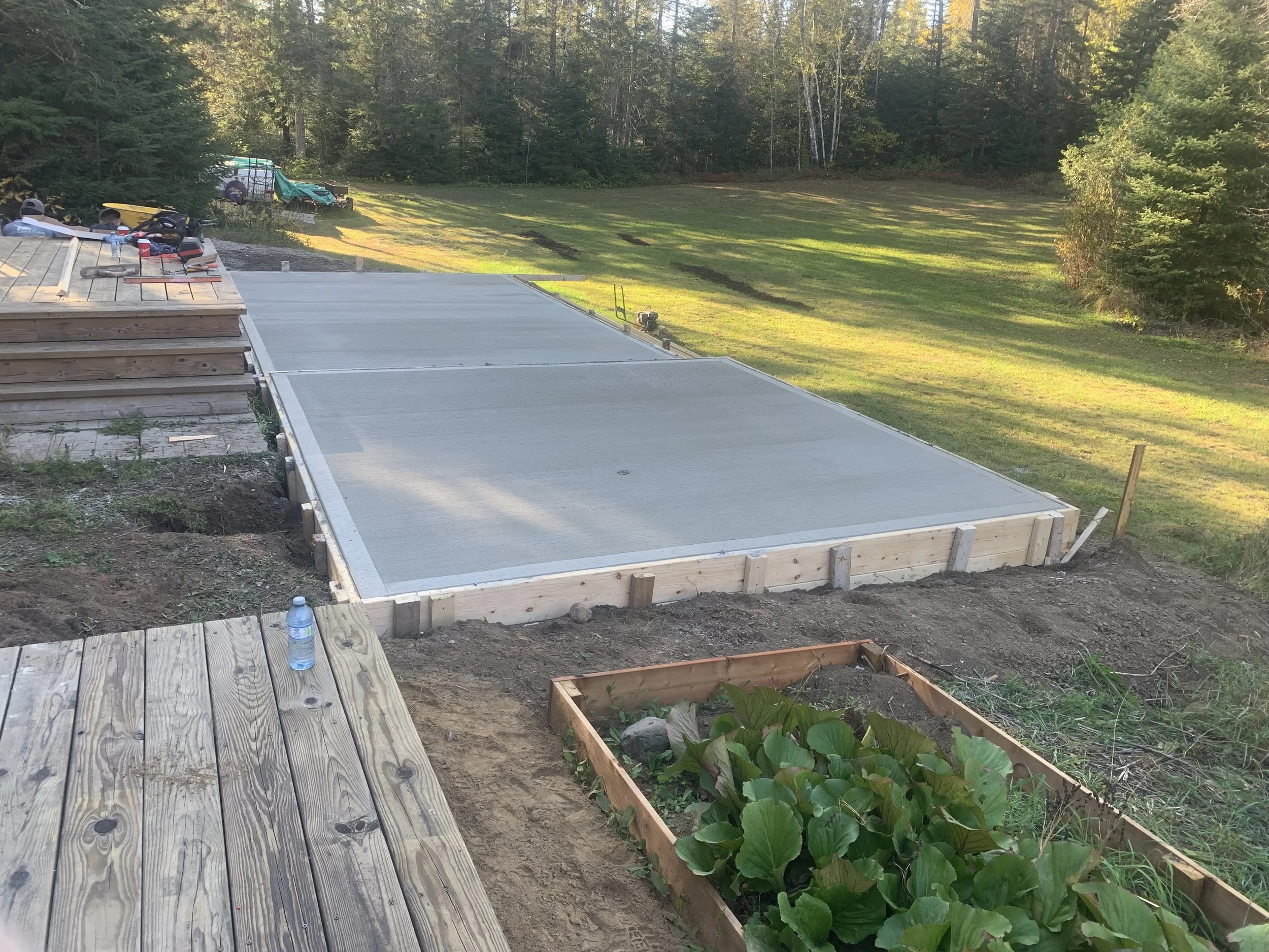 Construction site showing a freshly poured concrete slab foundation, surrounded by wooden framing and tools, with a grassy field and trees in the background.