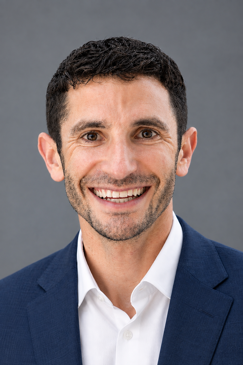 A smiling man with short, curly dark hair, a beard, and wearing a white shirt with a dark blue suit jacket, posed against a gray background.
