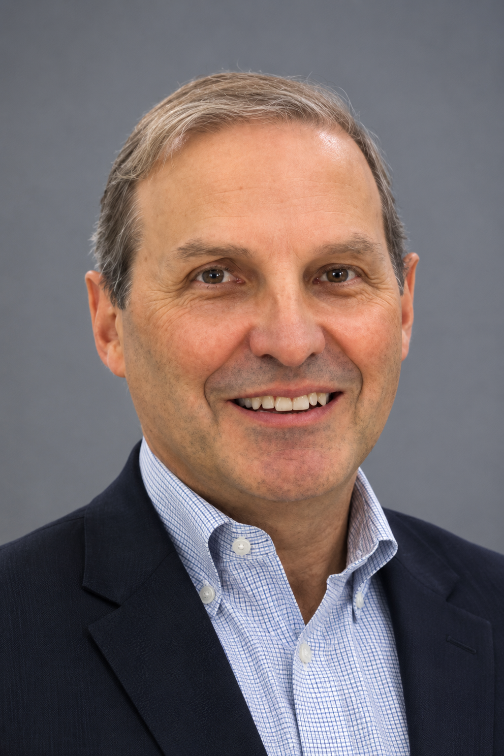 Close-up of a middle-aged man with short, light brown hair, wearing a dark suit jacket and a light blue checkered shirt, smiling against a gray background.