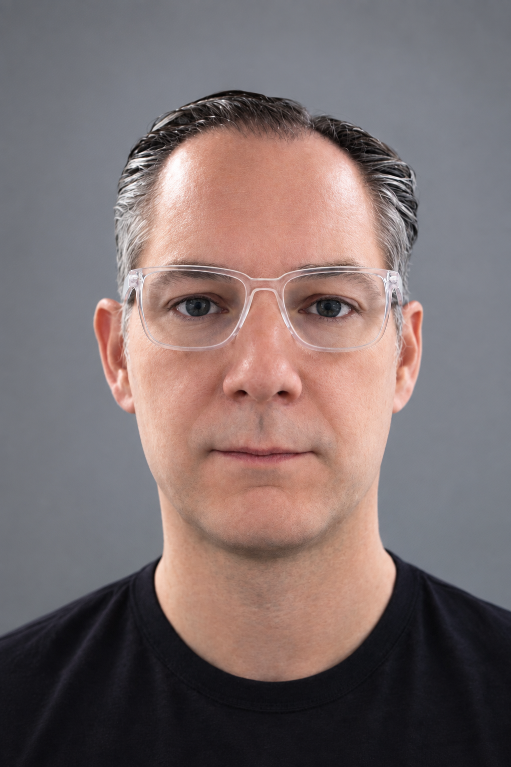 A man with light skin, dark gray hair, blue eyes, wearing clear glasses, and a black T-shirt, looking directly at the camera against a gray background.