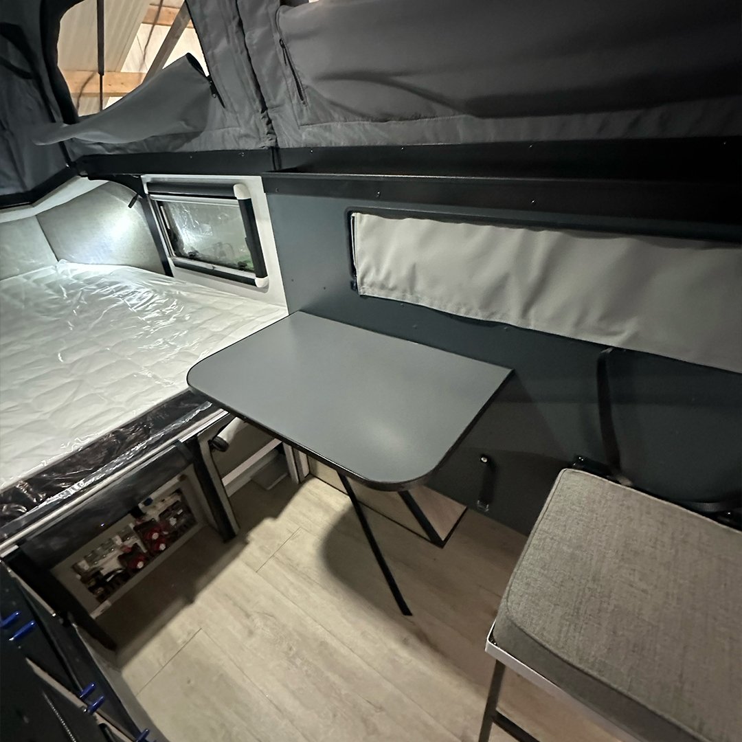 Interior of a camper van showing a small bed, a foldable table, and seating area, with storage cabinets and windows.