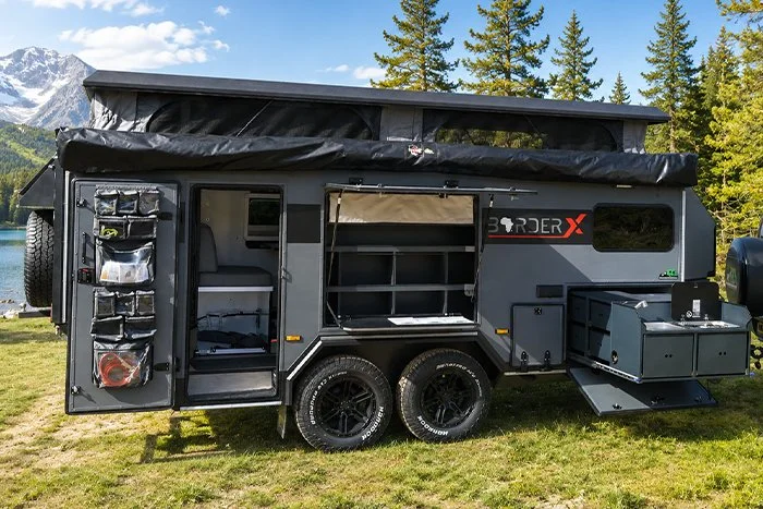 A black BorderX off-road camper trailer with storage compartments and an extended awning, set in a grassy area with trees and mountains in the background.