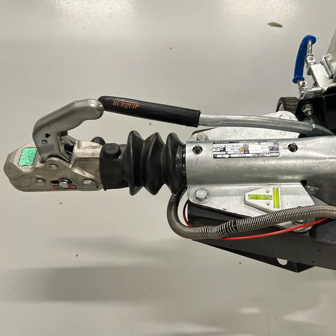 Close-up of a mechanical front axle assembly with a black and silver suspension arm, rubber boot, metal brackets, and electrical wires.