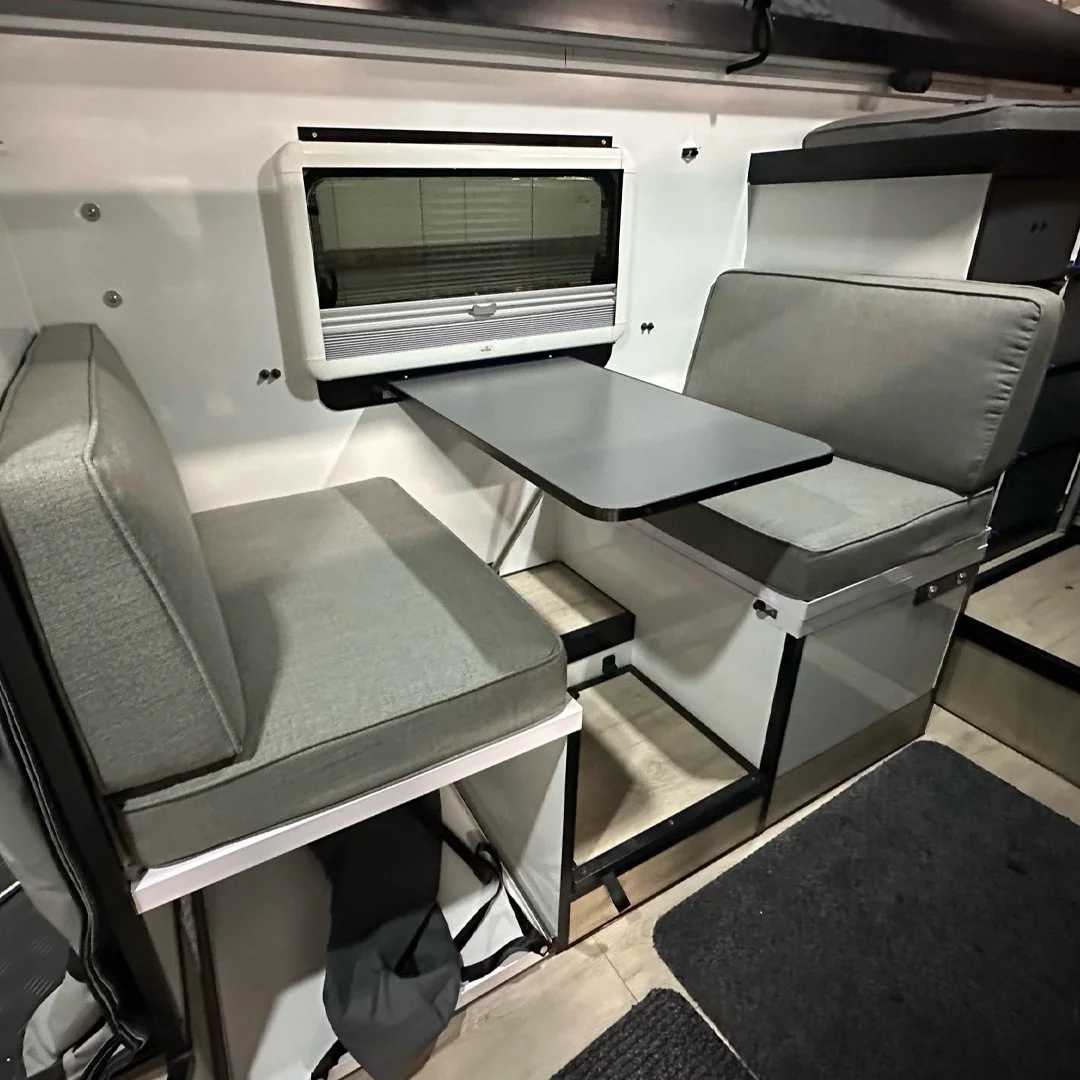The image shows a compact seating area inside a vehicle or small space, featuring two gray upholstered bench seats facing each other, a small rectangular table between them, and a window on the wall behind the table.