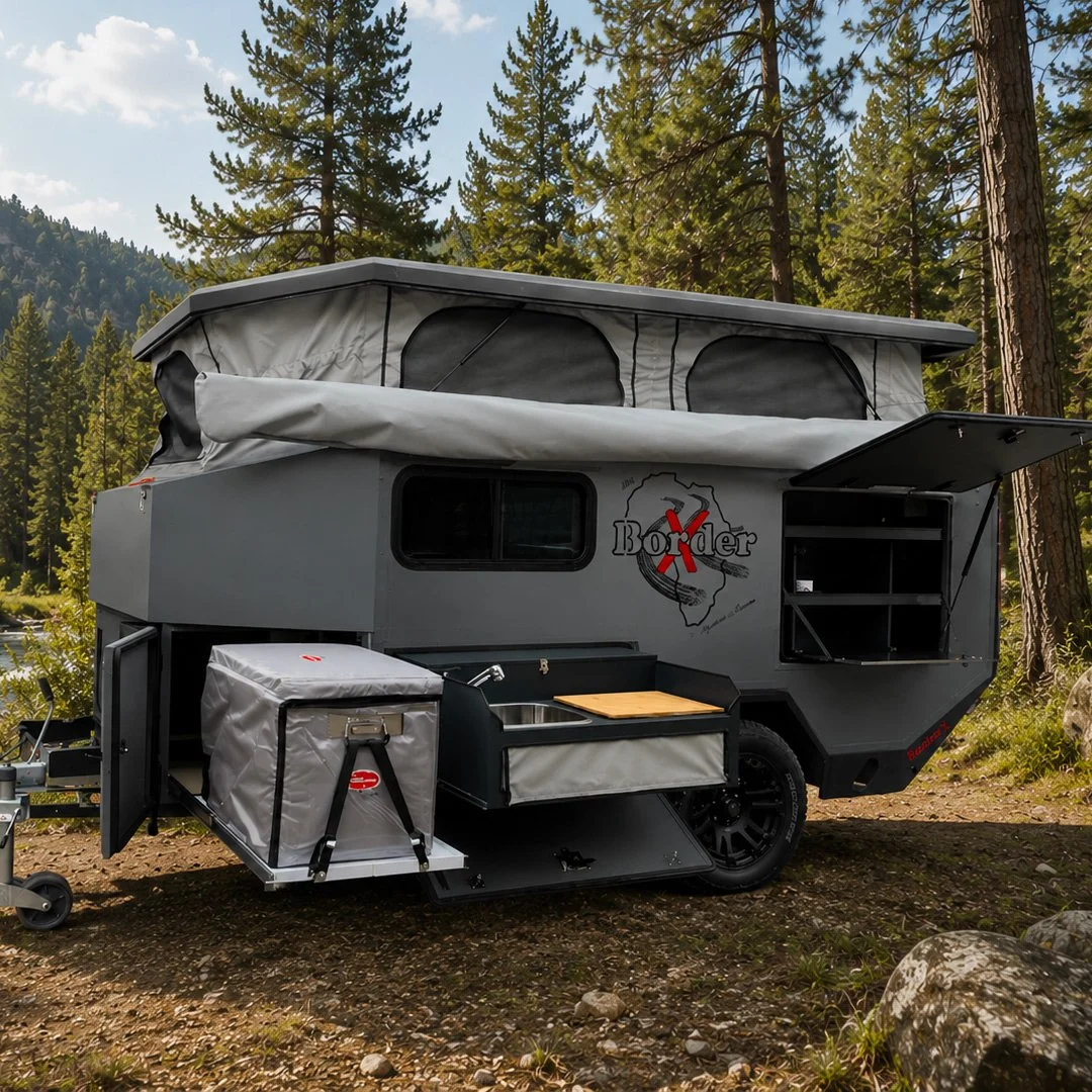 Grey off-road BorderX camper trailer with a pop-up tent and outdoor kitchen setup, positioned in a forested area with tall pine trees.