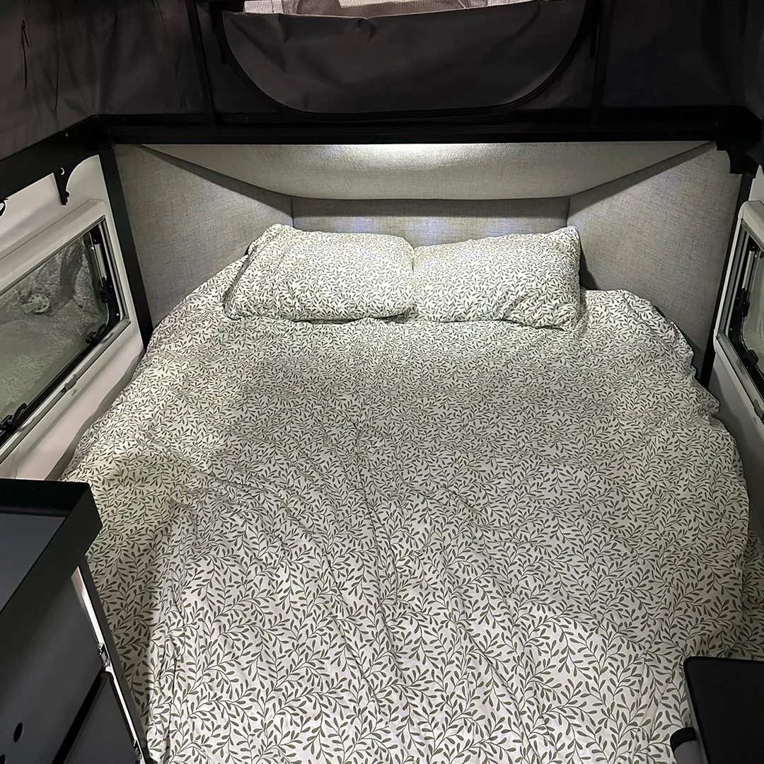 A neatly made bed inside a camper trailer with a patterned bedspread and pillows, surrounded by windows and interior walls.