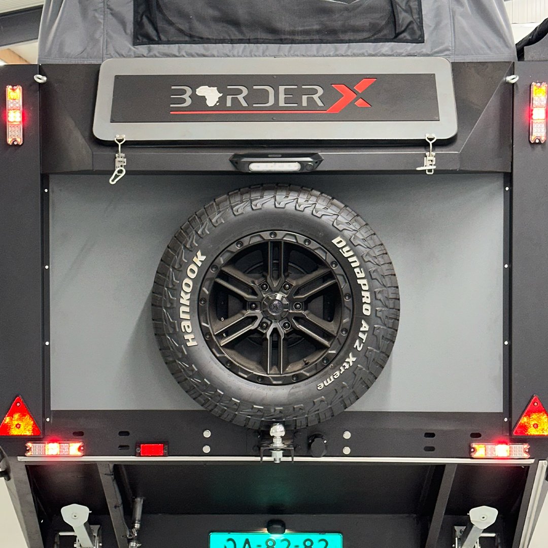 The back of an off-road utility trailer with a spare tire mounted on the center. The tire brand is Hankook, model is Dynapro A/T2. There is a logo labeled "BORDER X" above the tire, with a map of Africa inside the letter "O". The trailer has red and amber lights on the sides and a license plate at the bottom.