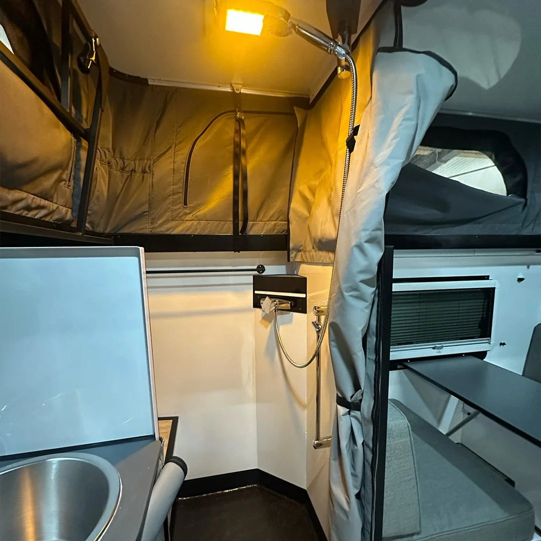 Interior of a compact camper or mobile living space with a small kitchen sink, a shower with a handheld showerhead, and a sleeping area with a raised bed and window, illuminated by a yellowish light.