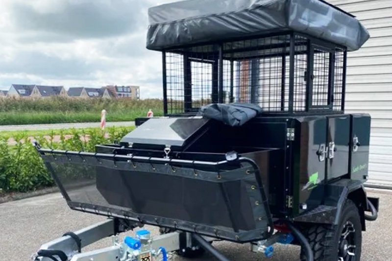 Off-road utility trailer with metal cage, black exterior, and storage compartments, parked outdoors.