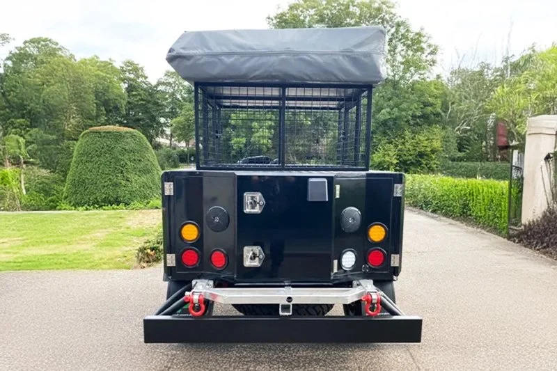 Rear view of a black utility vehicle with a metal cage and lights, parked on a driveway surrounded by greenery.