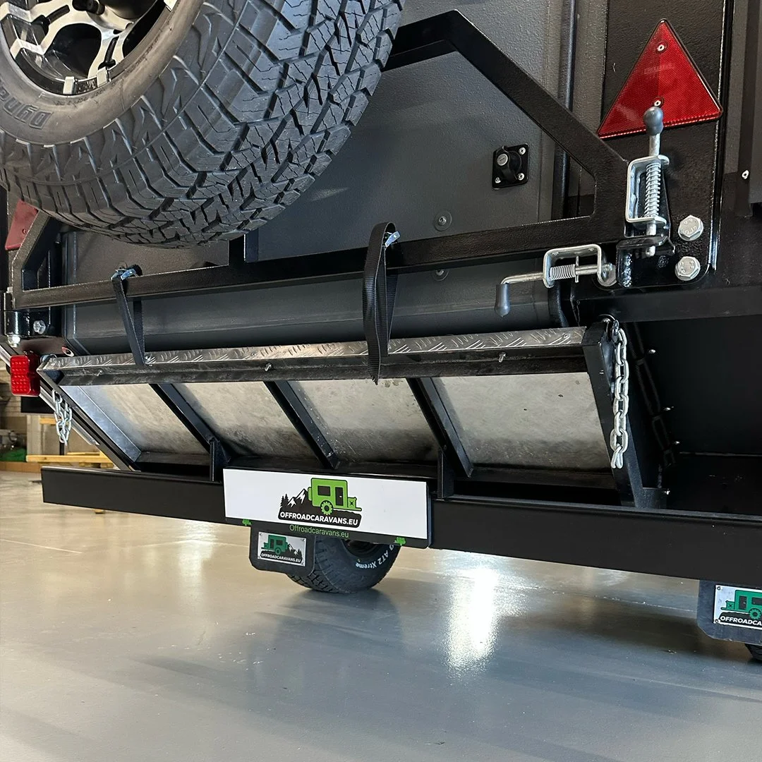 A black off-road camper trailer with a spare tire mounted on top, a trailor hitch with chains, a red triangle reflector, and a metal step at the back, parked on a smooth indoor floor.