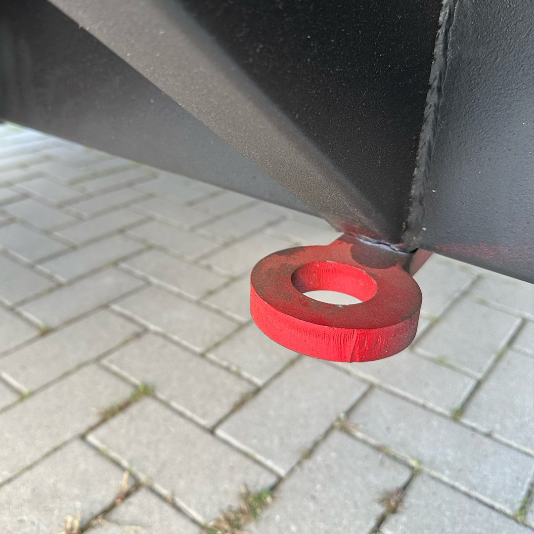 A red metal eye bolt attached to a black metal surface, with a paved surface of brick pavers underneath.