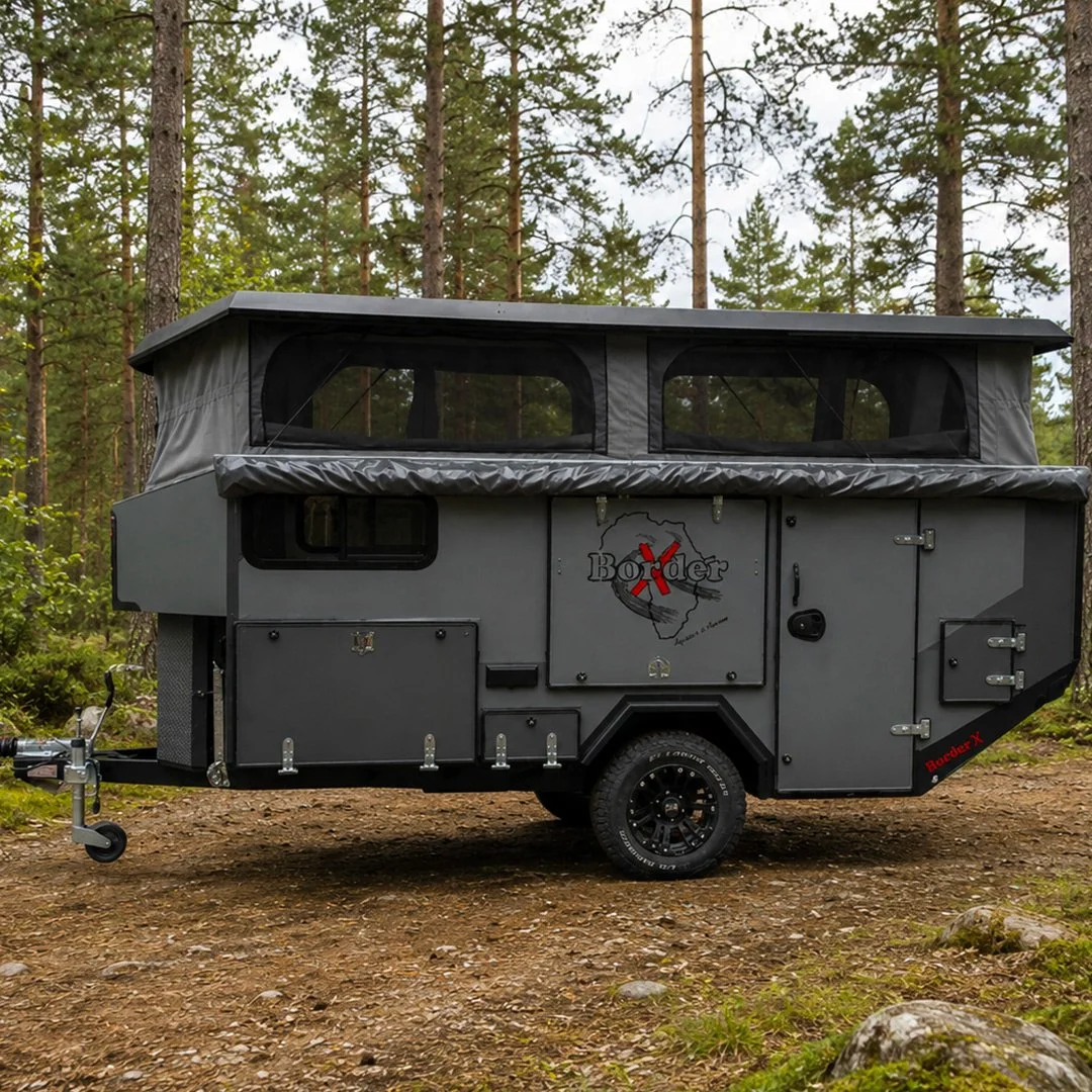A gray off-road BorderX camper trailer with a canopy roof, parked on dirt in a wooded forest area.