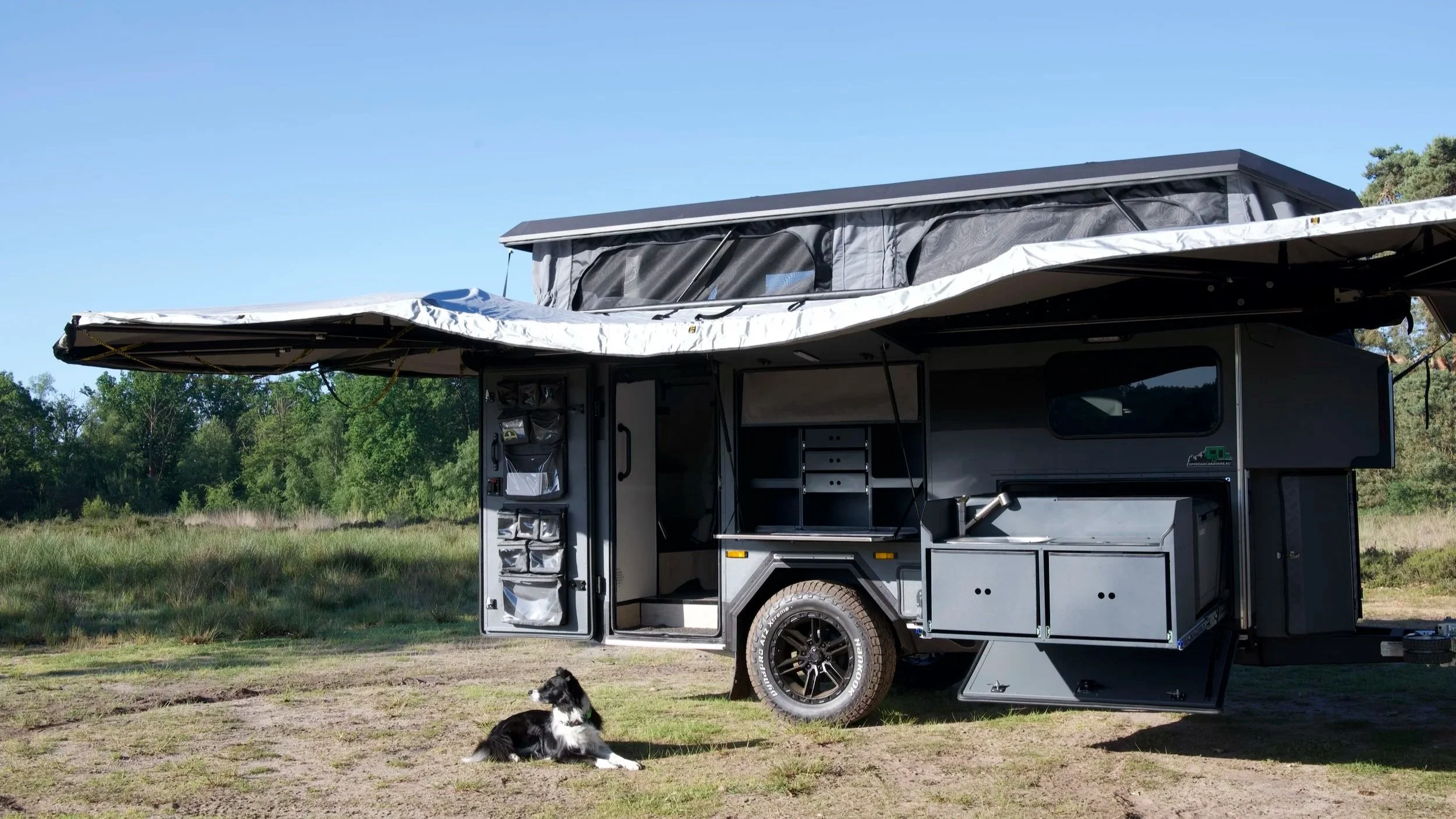 A black off-road BorderX camper trailer with an extended pop-up roof, set up on a grassy field with green trees in the background. A black and white dog is sitting in the foreground near the trailer.