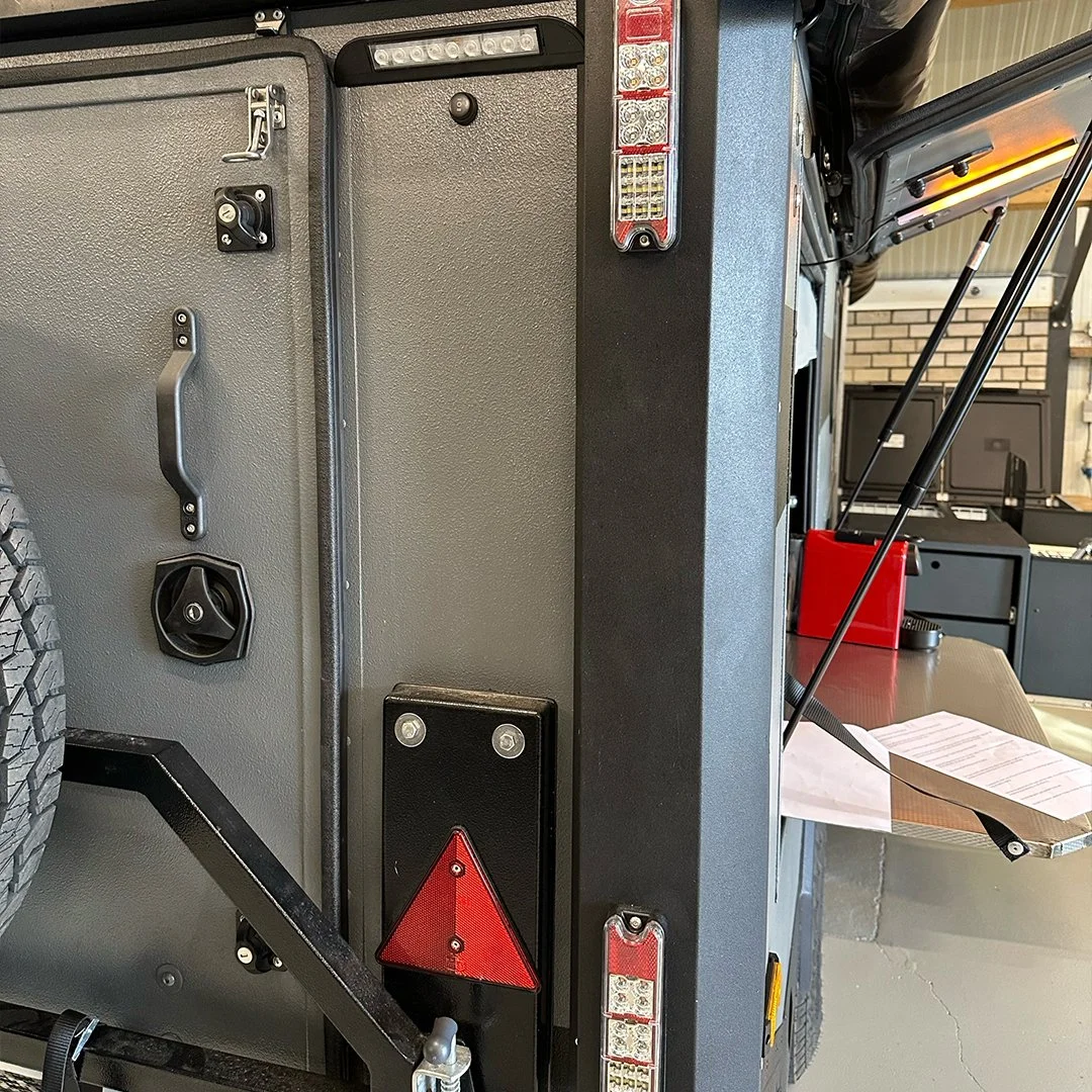 Close-up of the back of a utility vehicle, showing taillights, hand grips, and safety reflectors, with paper documents and a red box on a fold-out table.