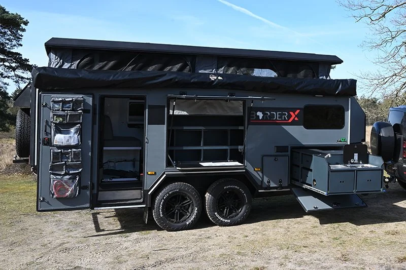 A gray off-road BorderX camper trailer with storage compartments, an awning, and rugged tires, parked on a patch of grass with trees in the background.