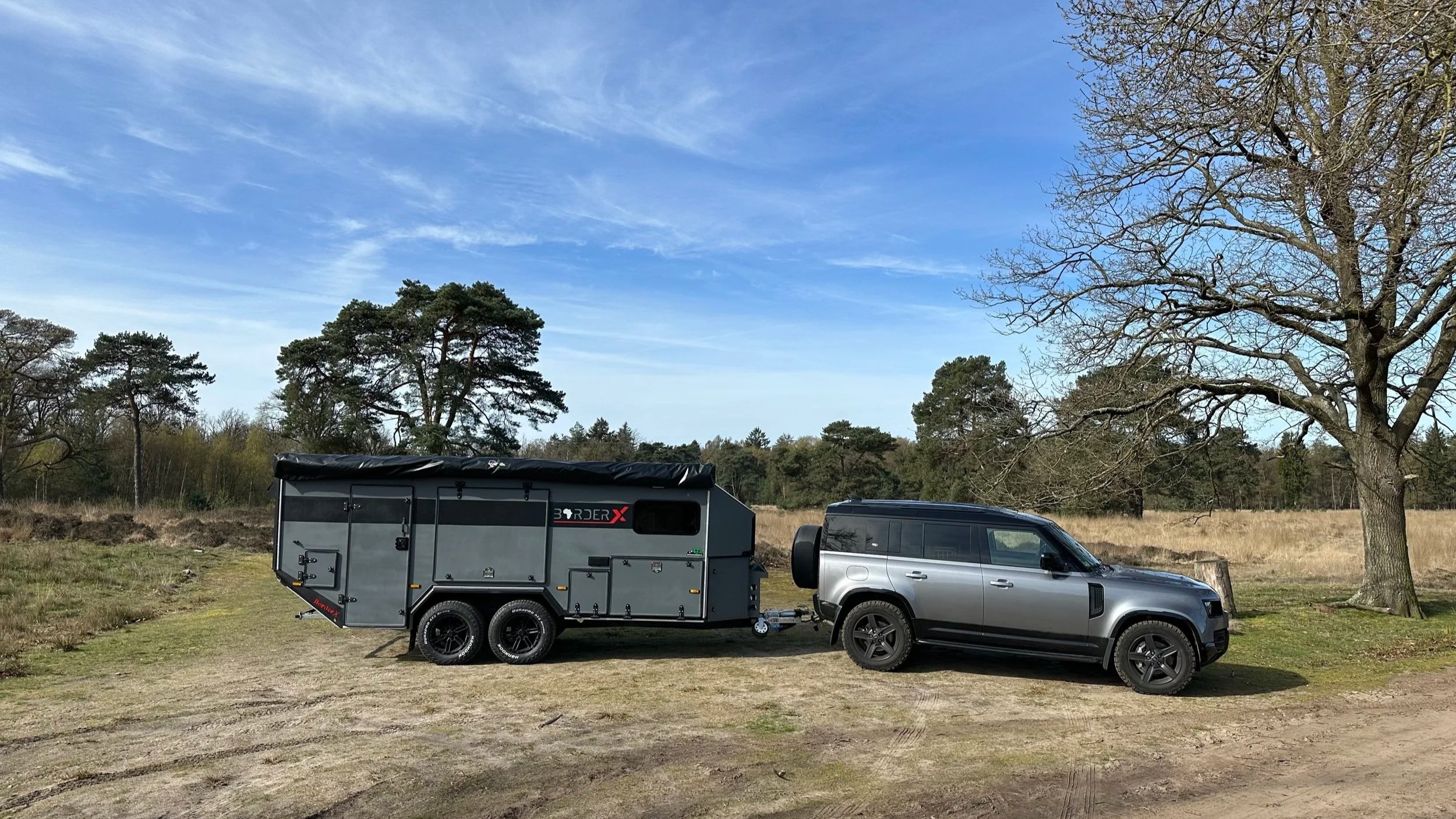 A silver SUV parked on a grassy field, connected to a gray BorderX camper trailer. There are trees with mostly bare branches and a blue sky with some clouds in the background.
